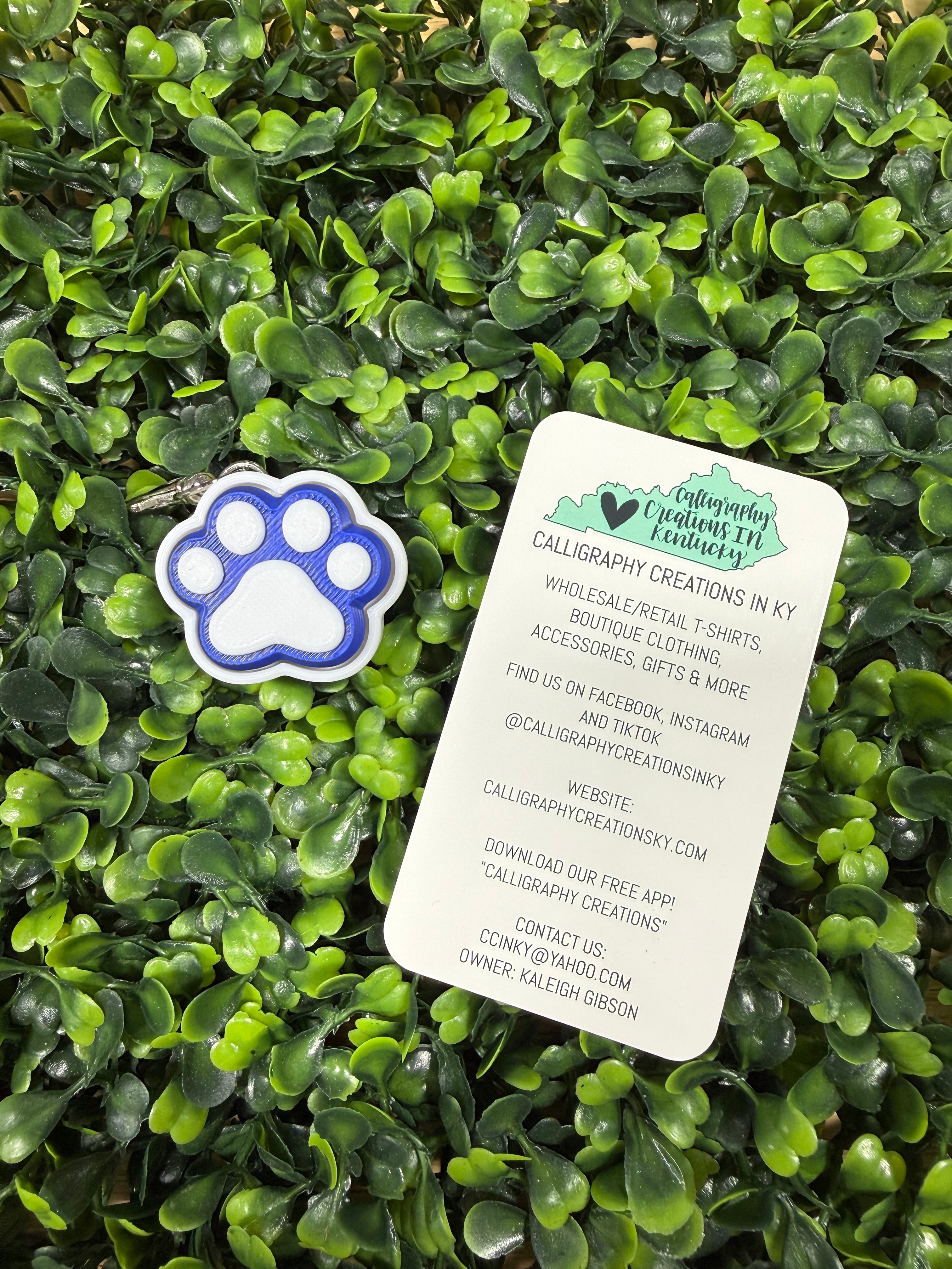 A small paw-shaped accessory with blue and white coloring lies on a white paper tag, both placed on dense green foliage.