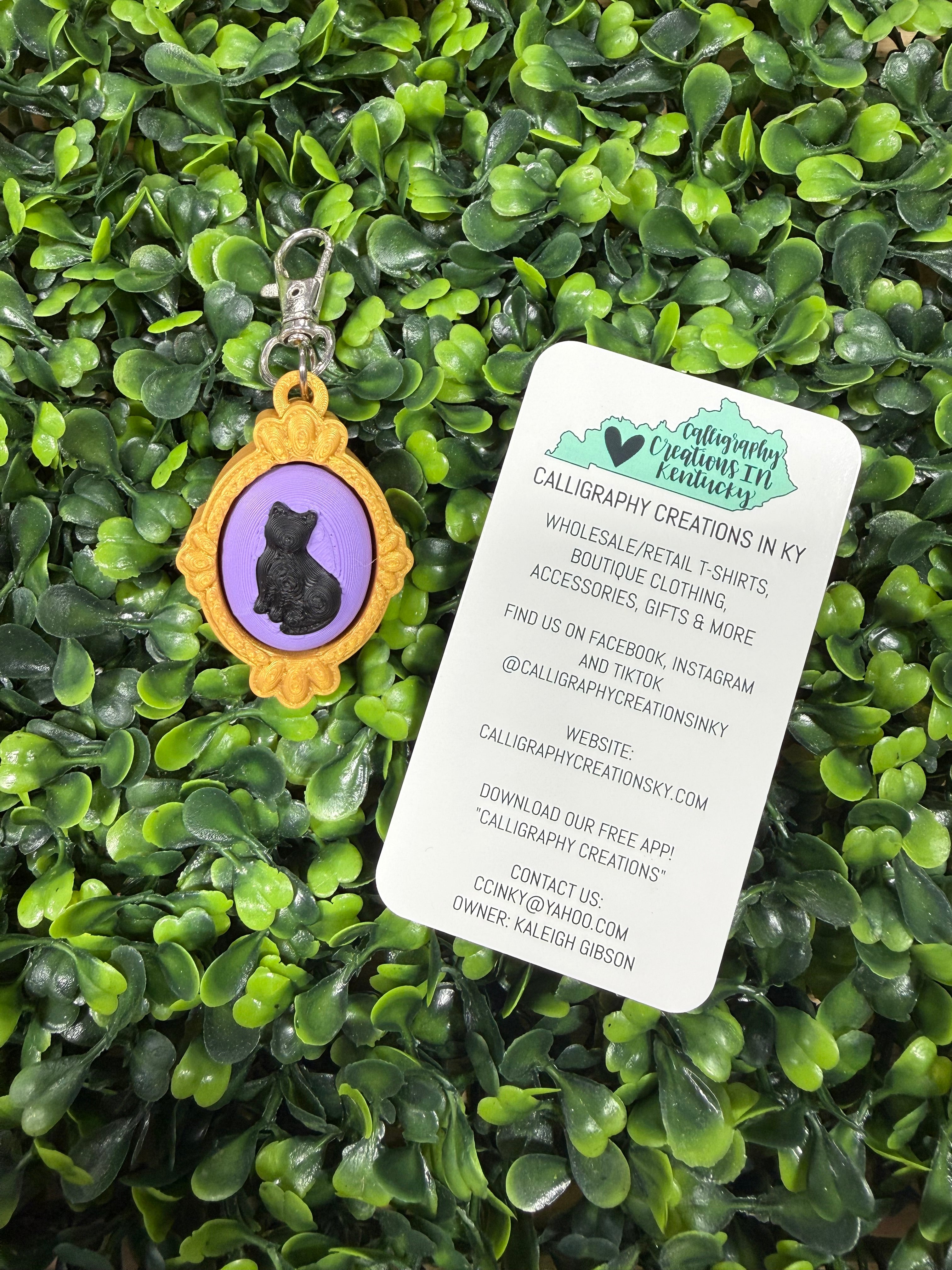A small oval pendant with a lavender-purple center and a gold-tone frame lies on green artificial foliage. A white paper tag with printed contact information is placed above it.