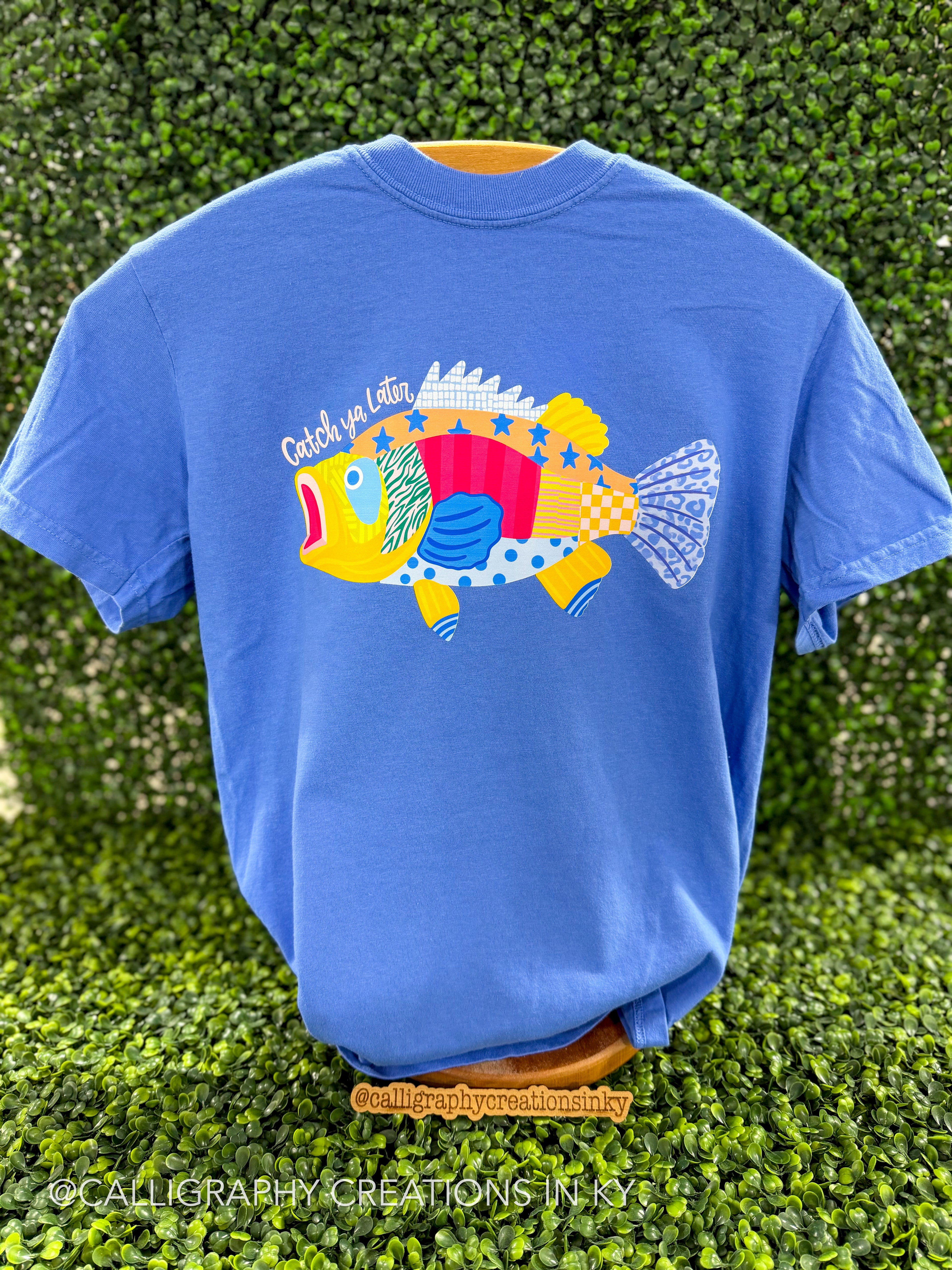 Blue shirt with patterned fish and the saying “Catch ya later” on a wooden stand 