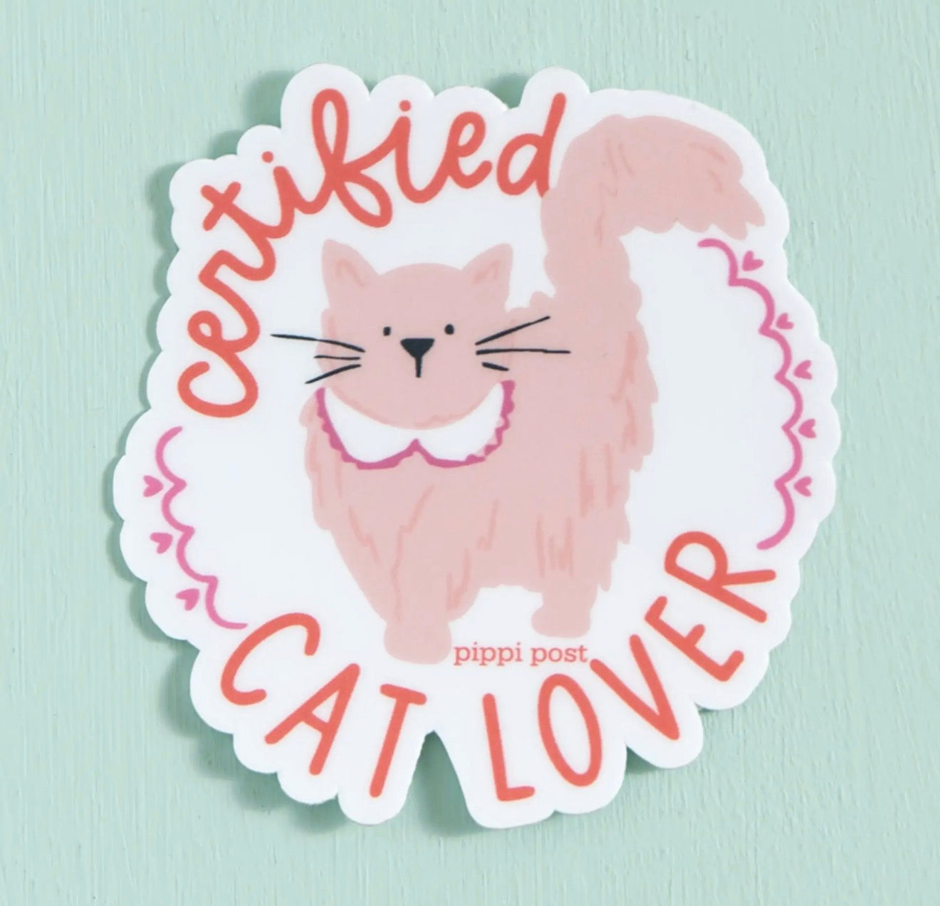 A die-cut sticker featuring a cute illustration of a fluffy pink cat wearing a white collar, surrounded by the text 'Certified Cat Lover' in coral pink lettering. The sticker is displayed on a light mint green background.