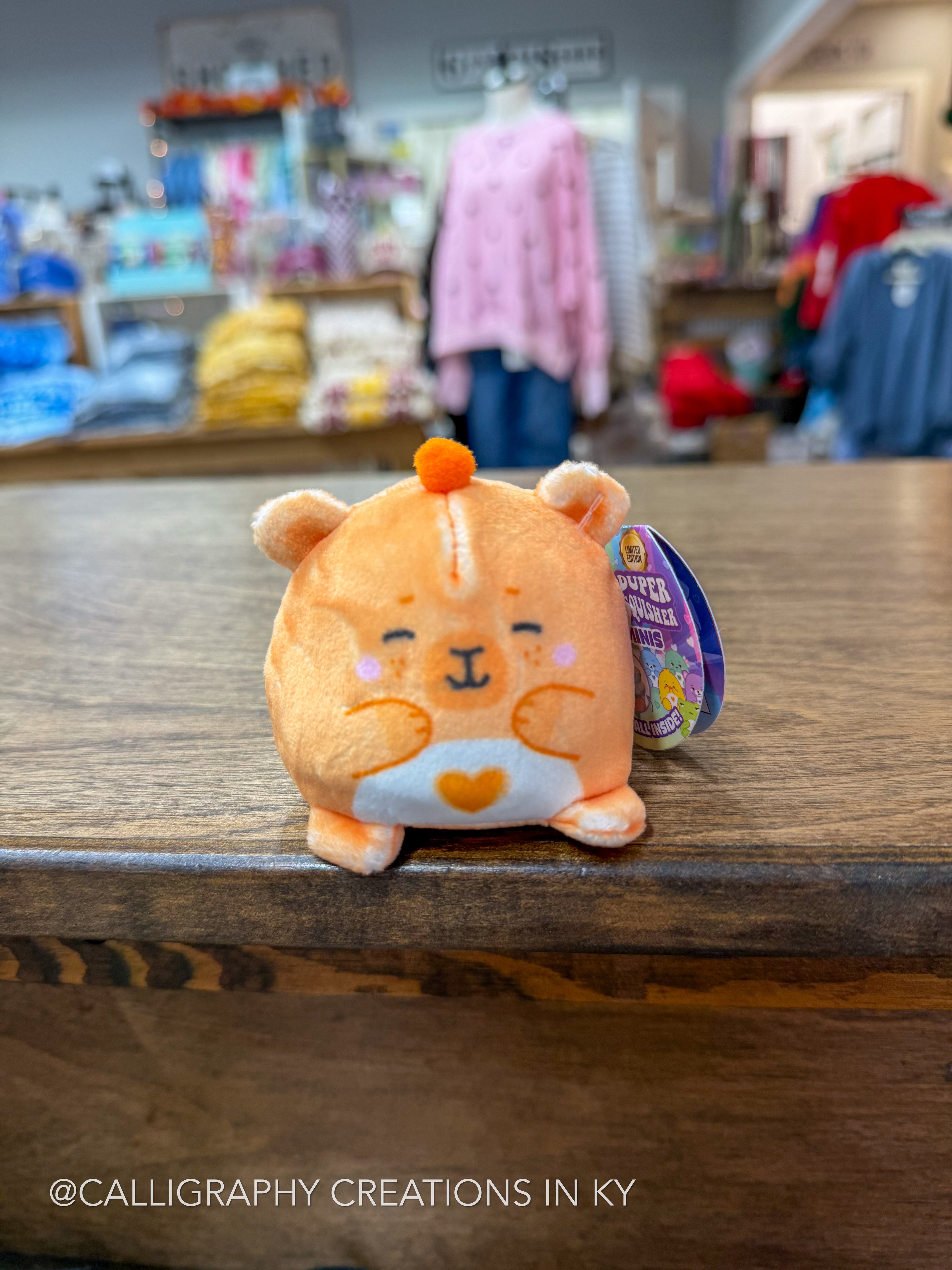 A small orange plush toy with a white belly and a heart-shaped chest detail sits on a wooden counter inside a store. The plush has a purple and yellow hangtag attached to its side.