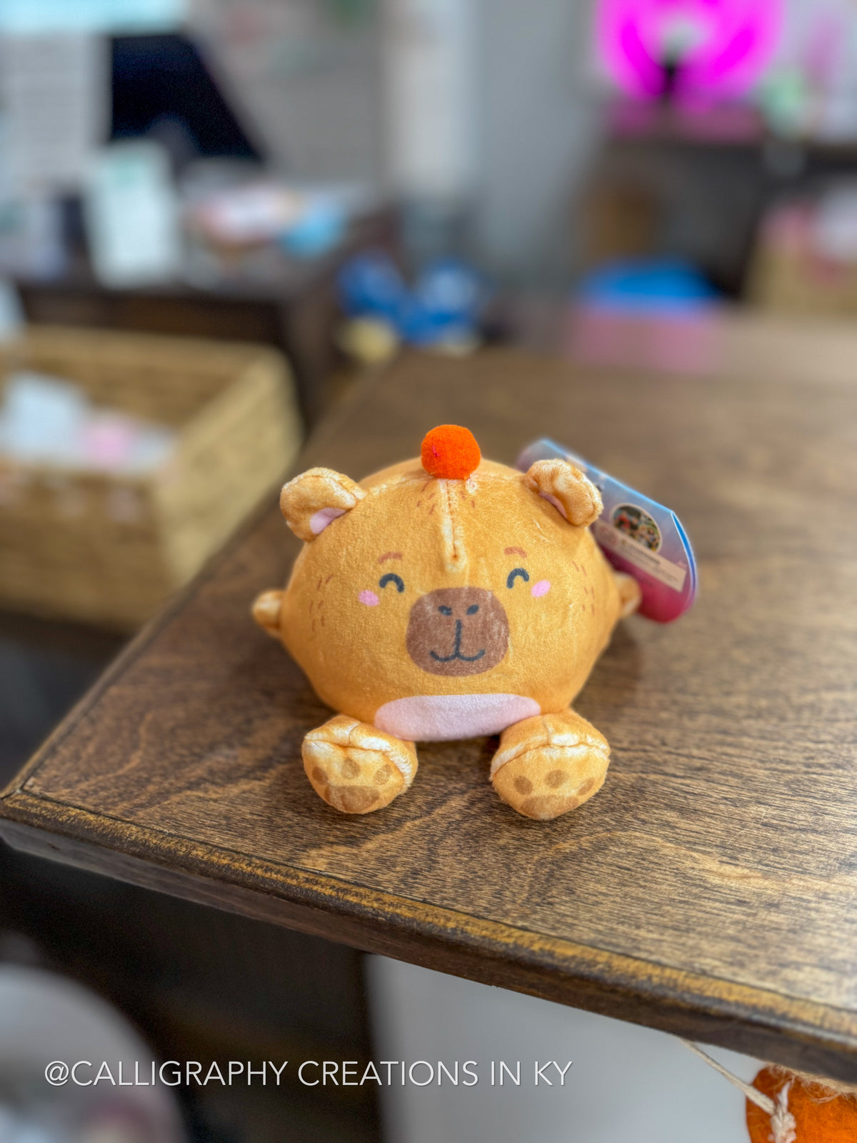 A small plush giraffe toy with a warm tan body, brown face and spots, pink inner ears, and an orange pom-pom, sitting on a wooden table. A hangtag is attached to the side, and the background is softly blurred.