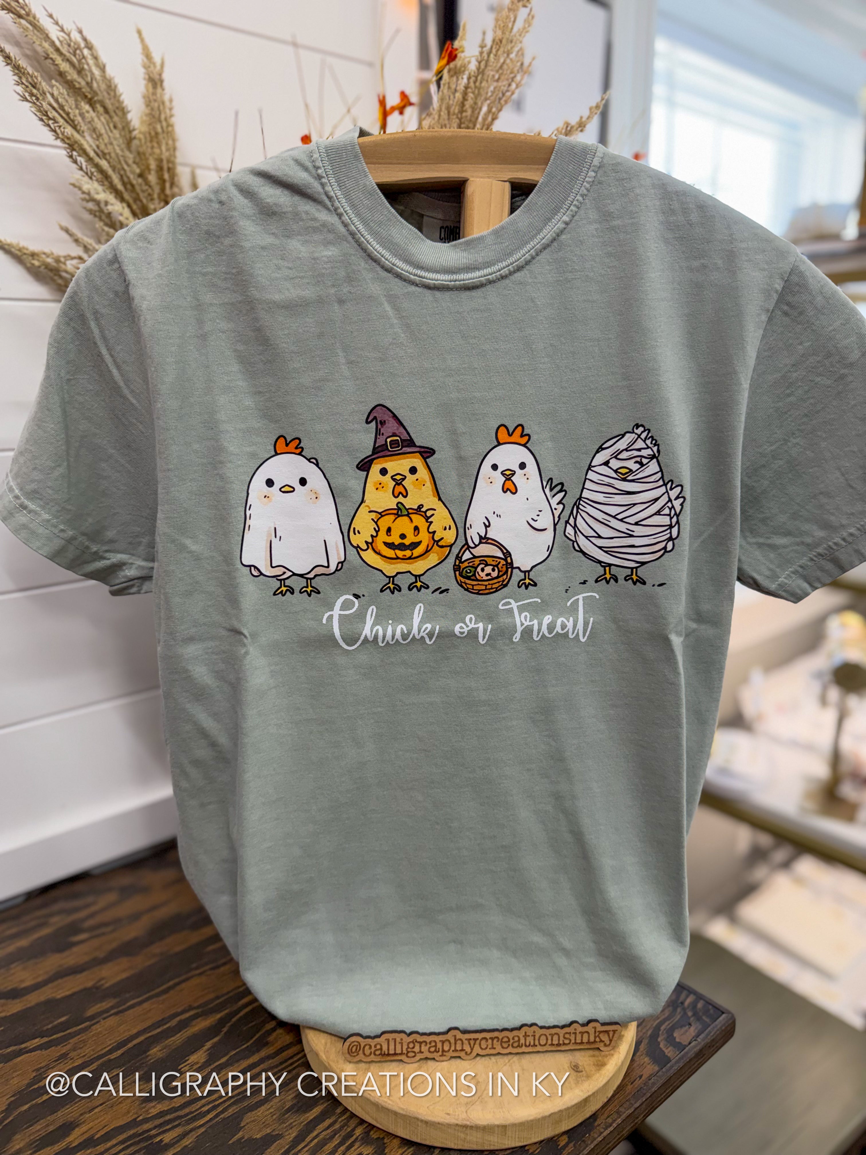 A sage green short-sleeve t-shirt on a wooden hanger, featuring a graphic of four cute cartoon chickens dressed in Halloween costumes with the text 'Chick or Treat' written below.