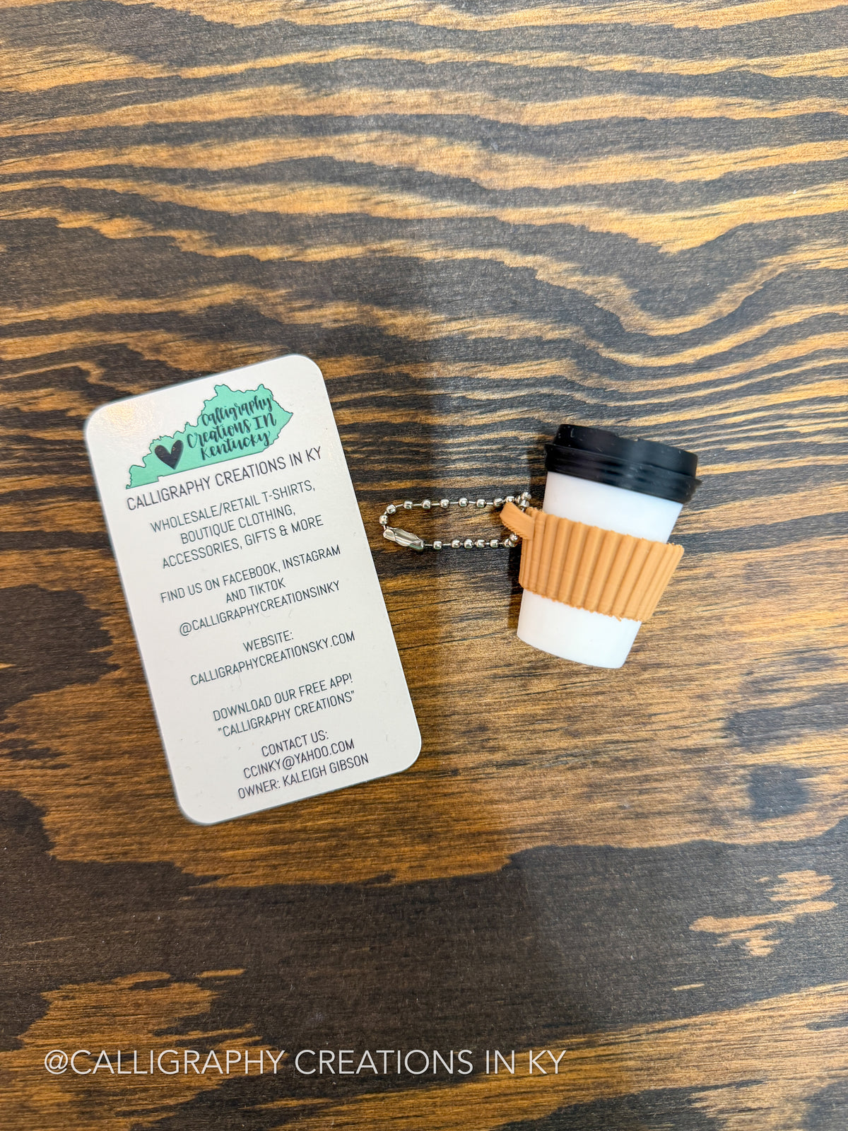 A coffee cup keychain charm with a black lid, white body, and tan sleeve, attached to a metal key ring, placed on a dark wood surface next to a white business card for Calligraphy Creations in KY.