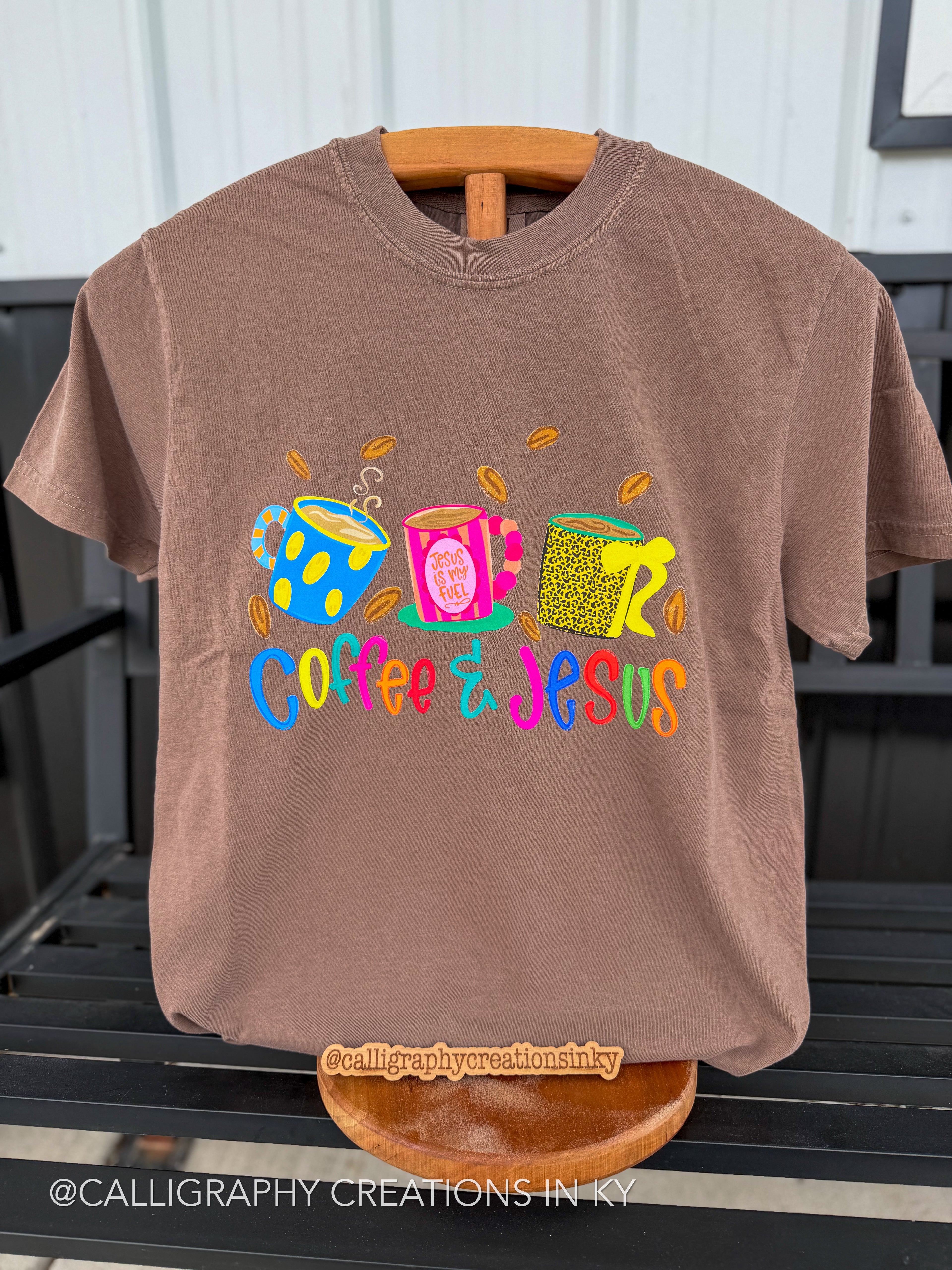 A brown short-sleeve t-shirt on a wooden hanger, featuring a colorful graphic of three coffee cups and the text 'Coffee Jesus' in vibrant multicolor lettering. The shirt is displayed on a black slatted bench.