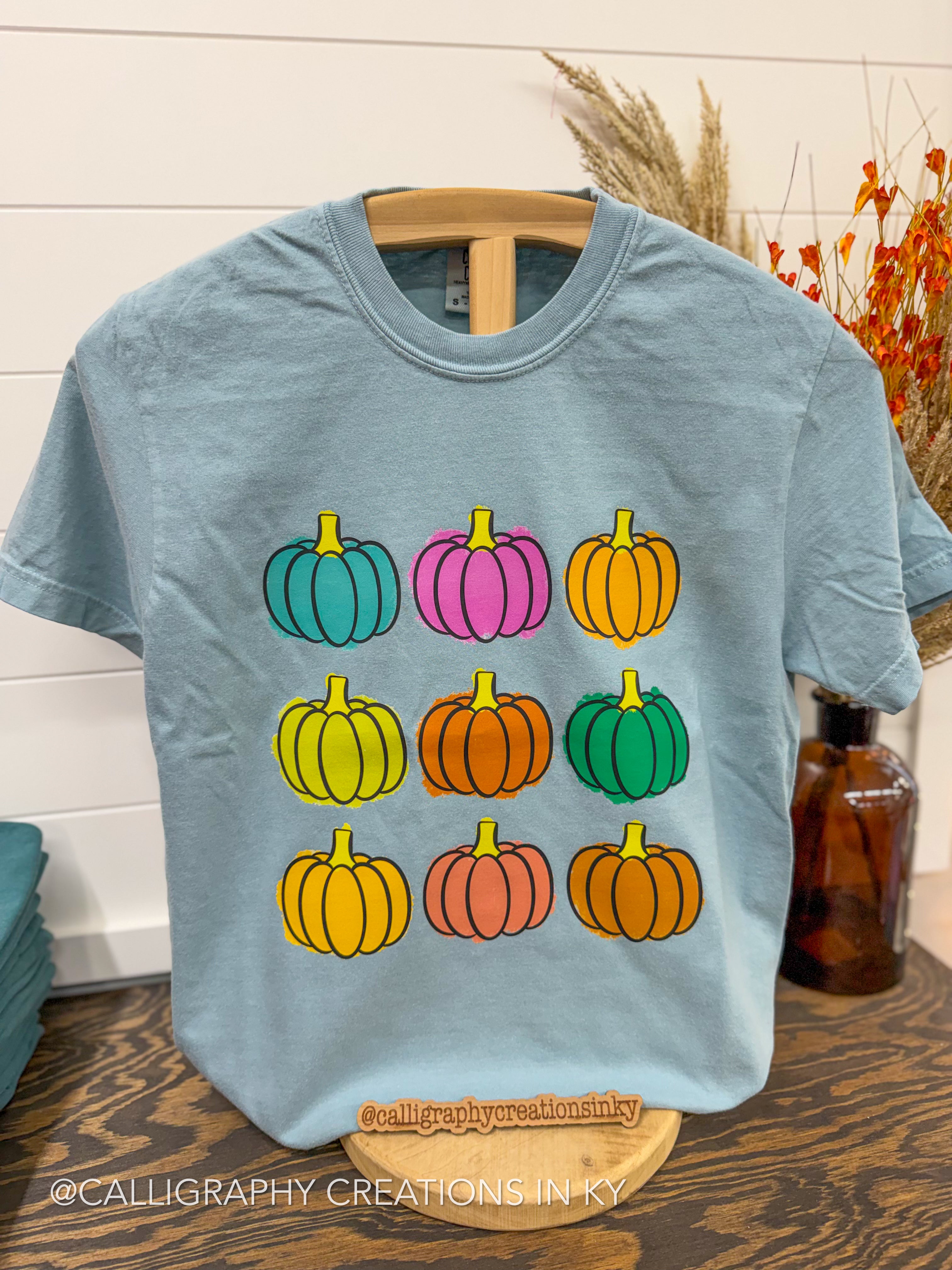 A light blue crewneck t-shirt on a wooden hanger, featuring a graphic of nine colorful pumpkins arranged in a 3x3 grid. The background shows a white shiplap wall, a wooden table, and dried grasses in a decorative vase.