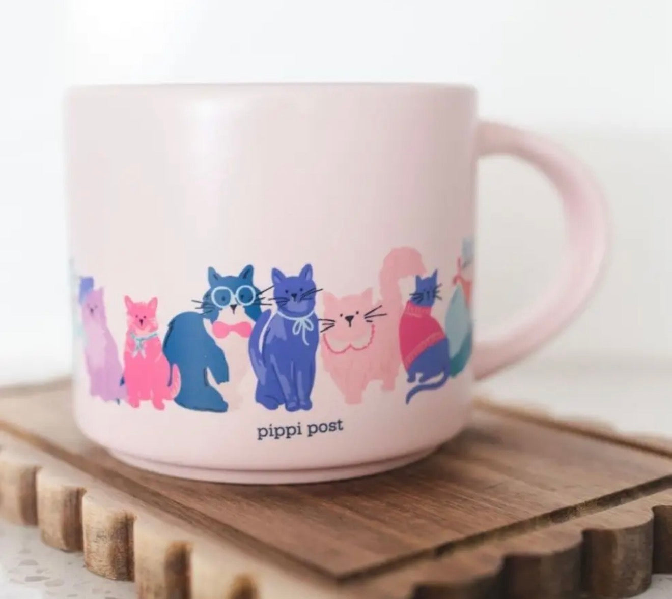 A light pink ceramic mug with a handle, decorated with a row of colorful cartoon cats and the text 'pippi post' near the bottom. The mug is resting on a wooden tray against a soft, out-of-focus white background.