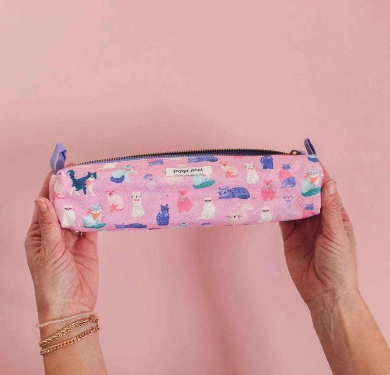 A pink zippered pencil case with a colorful cat pattern, held by two hands against a solid pink background.