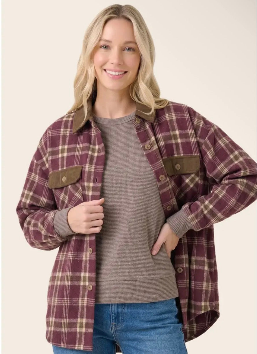 A woman modeling a burgundy and tan plaid button-up flannel shirt jacket over a taupe ribbed knit sweater and blue jeans, set against a solid light beige background.