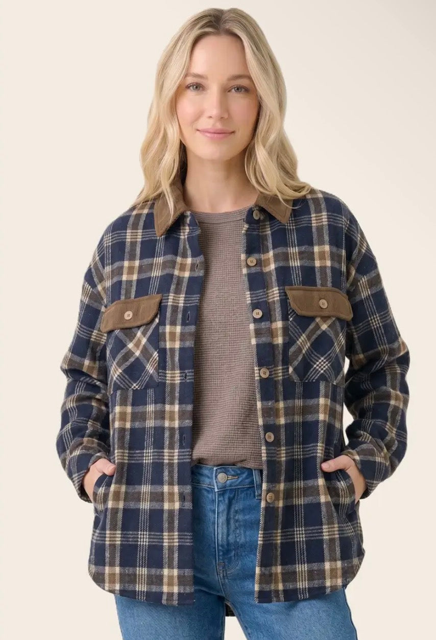 A woman modeling a navy blue plaid flannel shirt jacket with brown chest pockets, worn over a taupe knit top and blue jeans, against a solid light beige background.