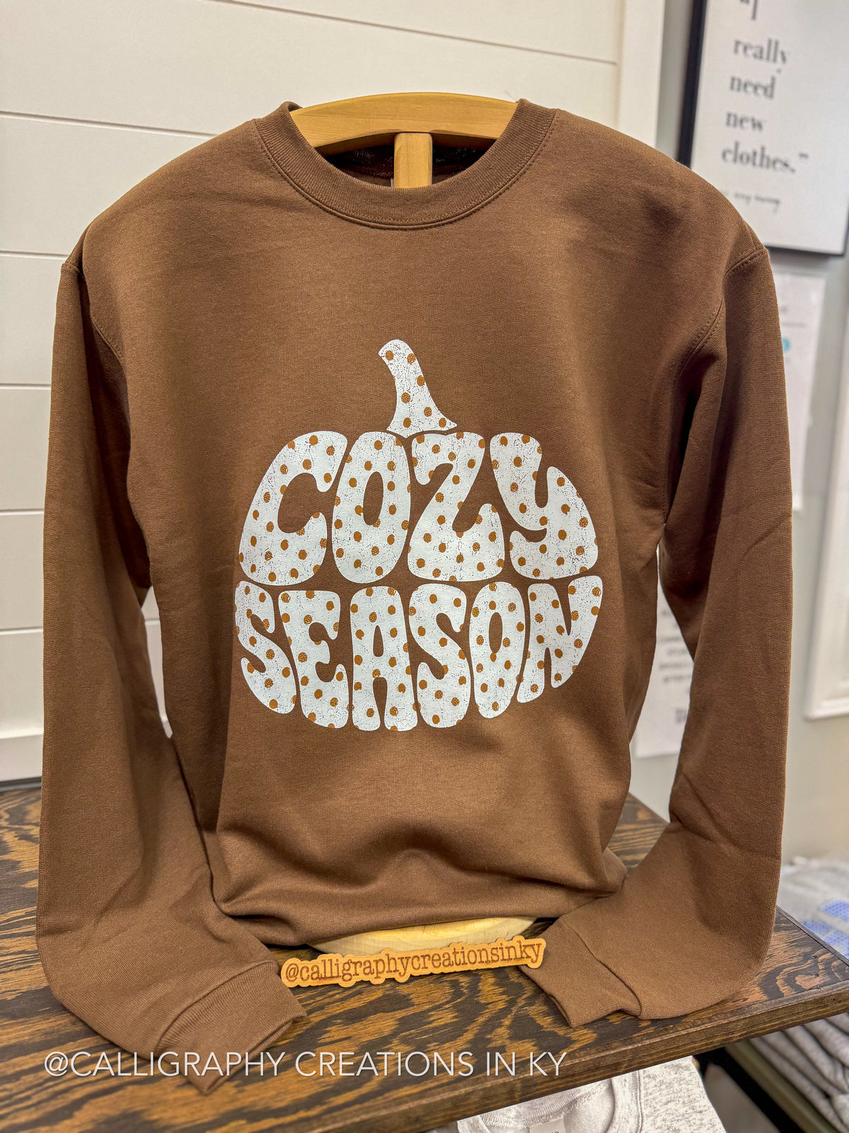 A brown crewneck sweatshirt on a wooden hanger, featuring a large graphic of a pumpkin made from the words 'COZY SEASON' in a white polka-dot pattern. The sweatshirt is displayed on a wooden table in a retail setting.