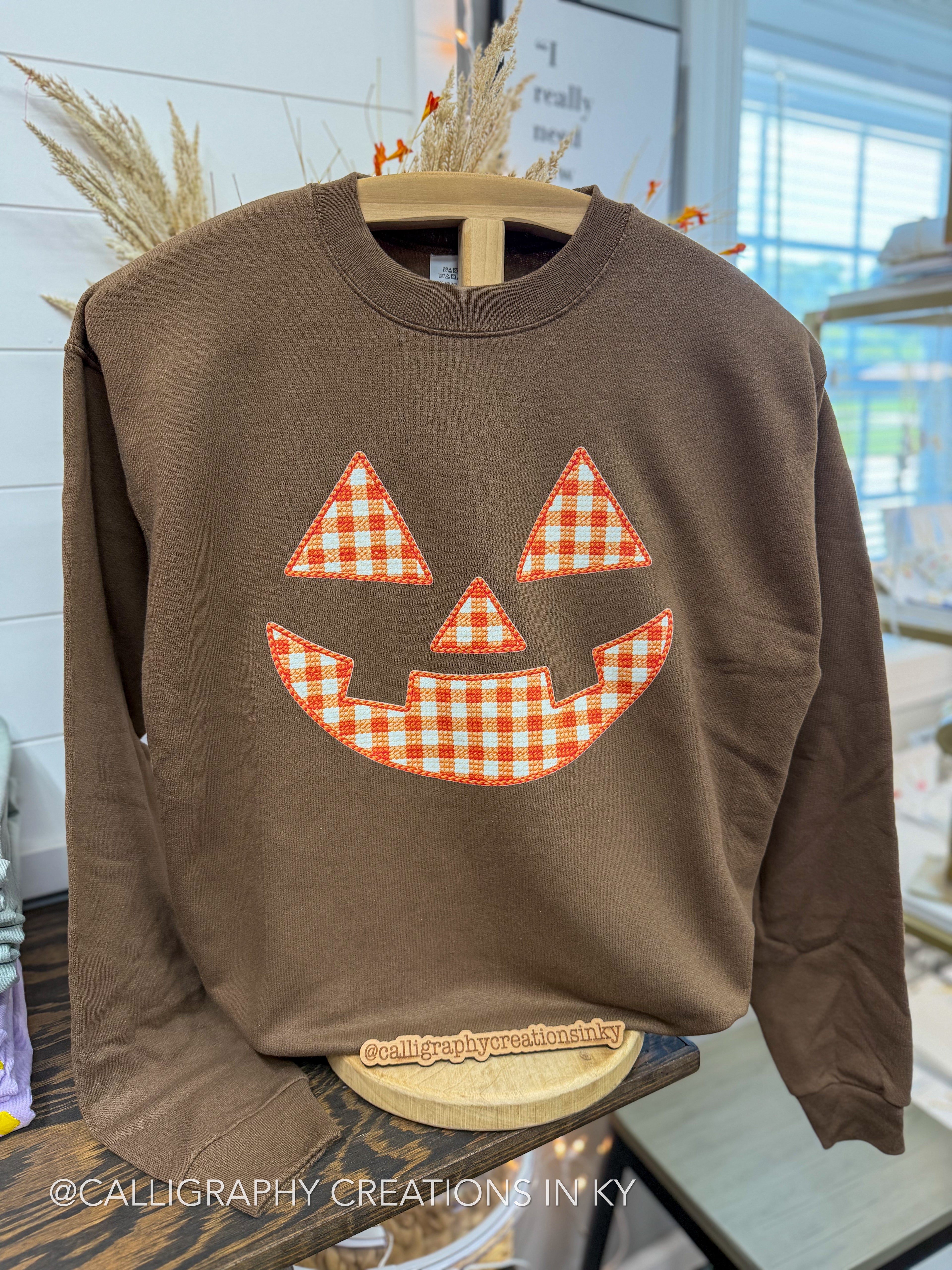 A brown crewneck sweatshirt featuring a pumpkin jack-o'-lantern face design in an orange and white gingham check pattern, displayed on a wooden hanger in a retail setting.