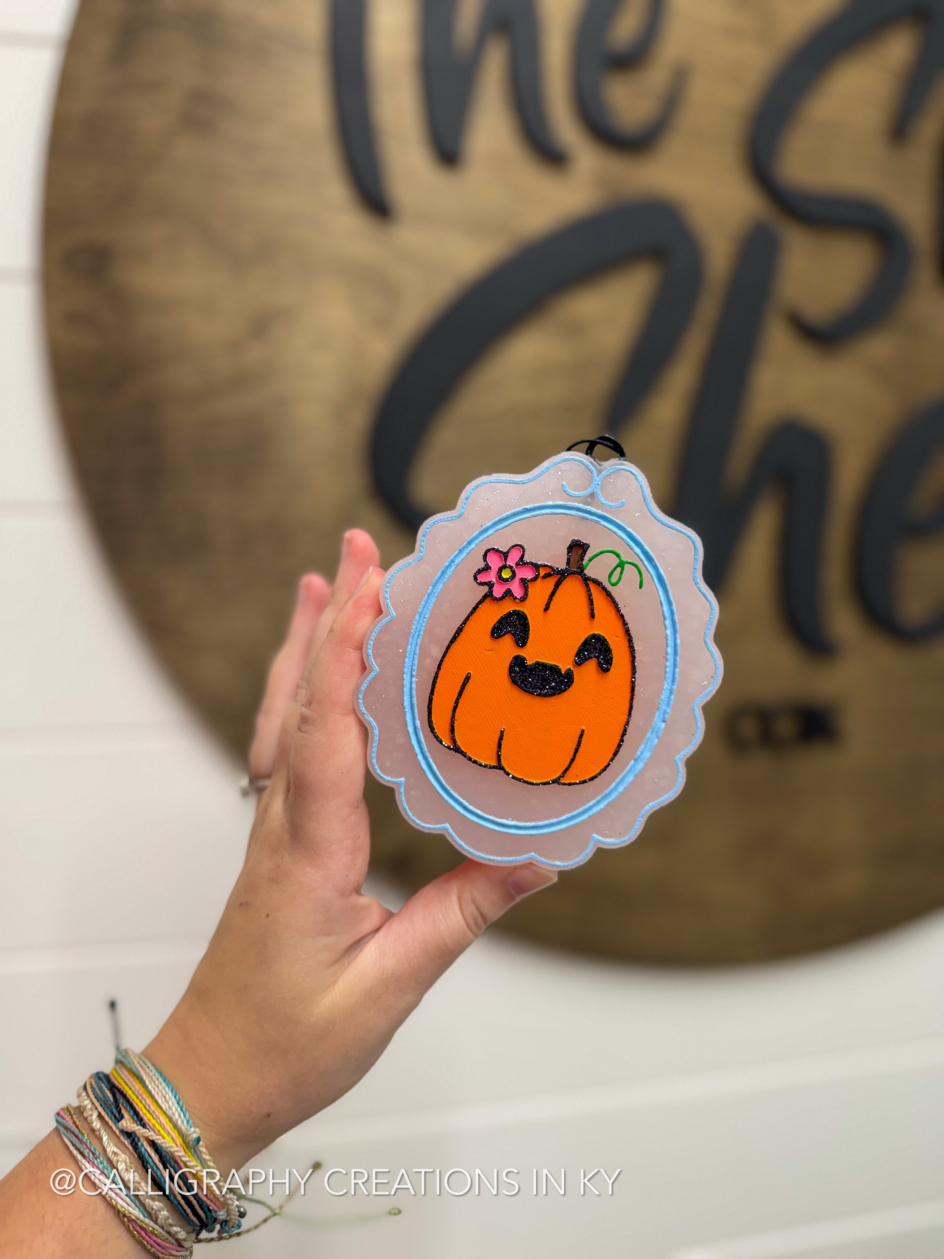 A hand holds a scalloped freshie featuring a cute orange pumpkin with a pink flower and green stem, set against a blurred rustic wooden sign background.