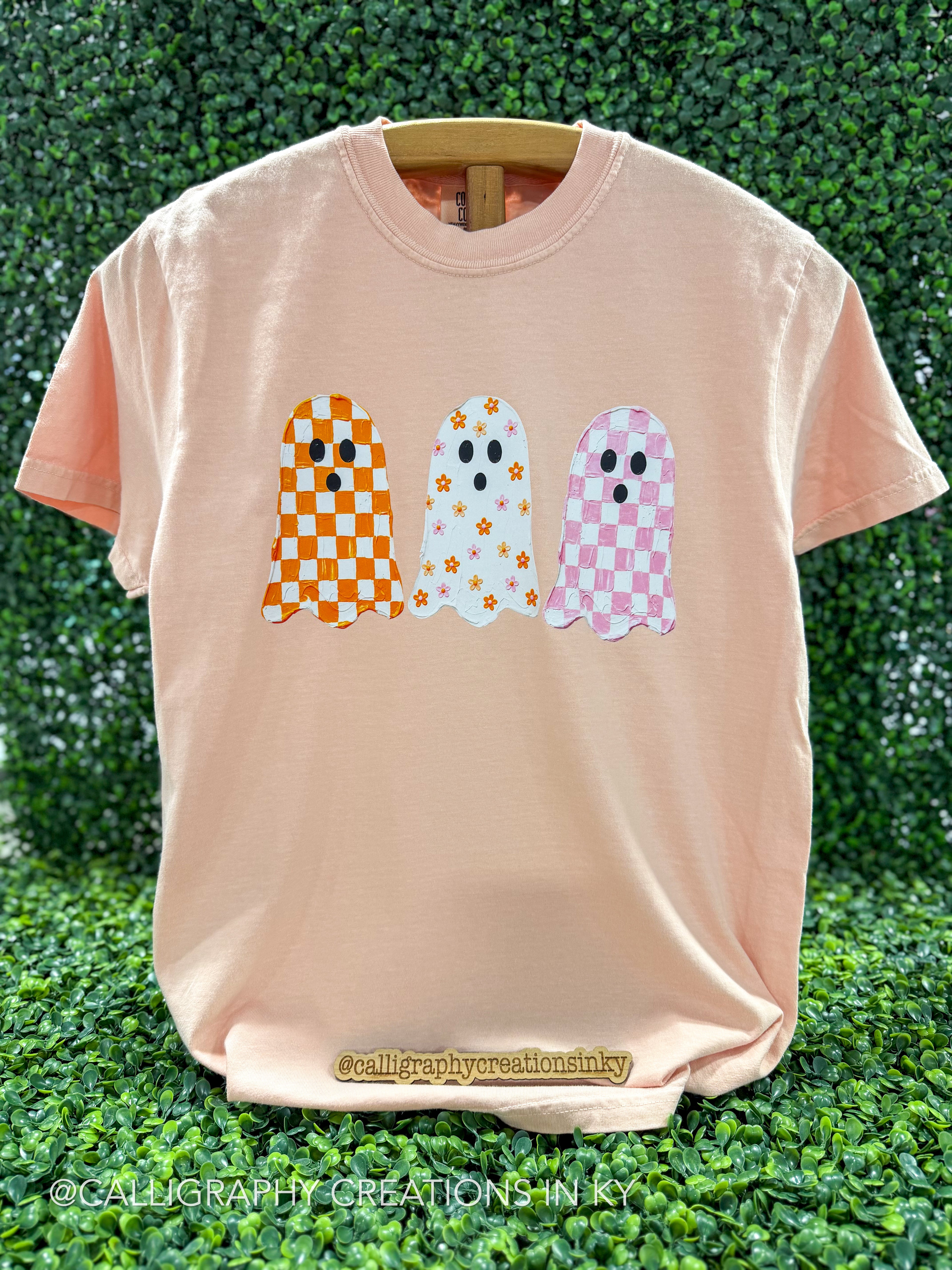 A peach-colored short-sleeve t-shirt with three cute ghost graphics on the front, displayed on a wooden hanger against a green leafy hedge background.