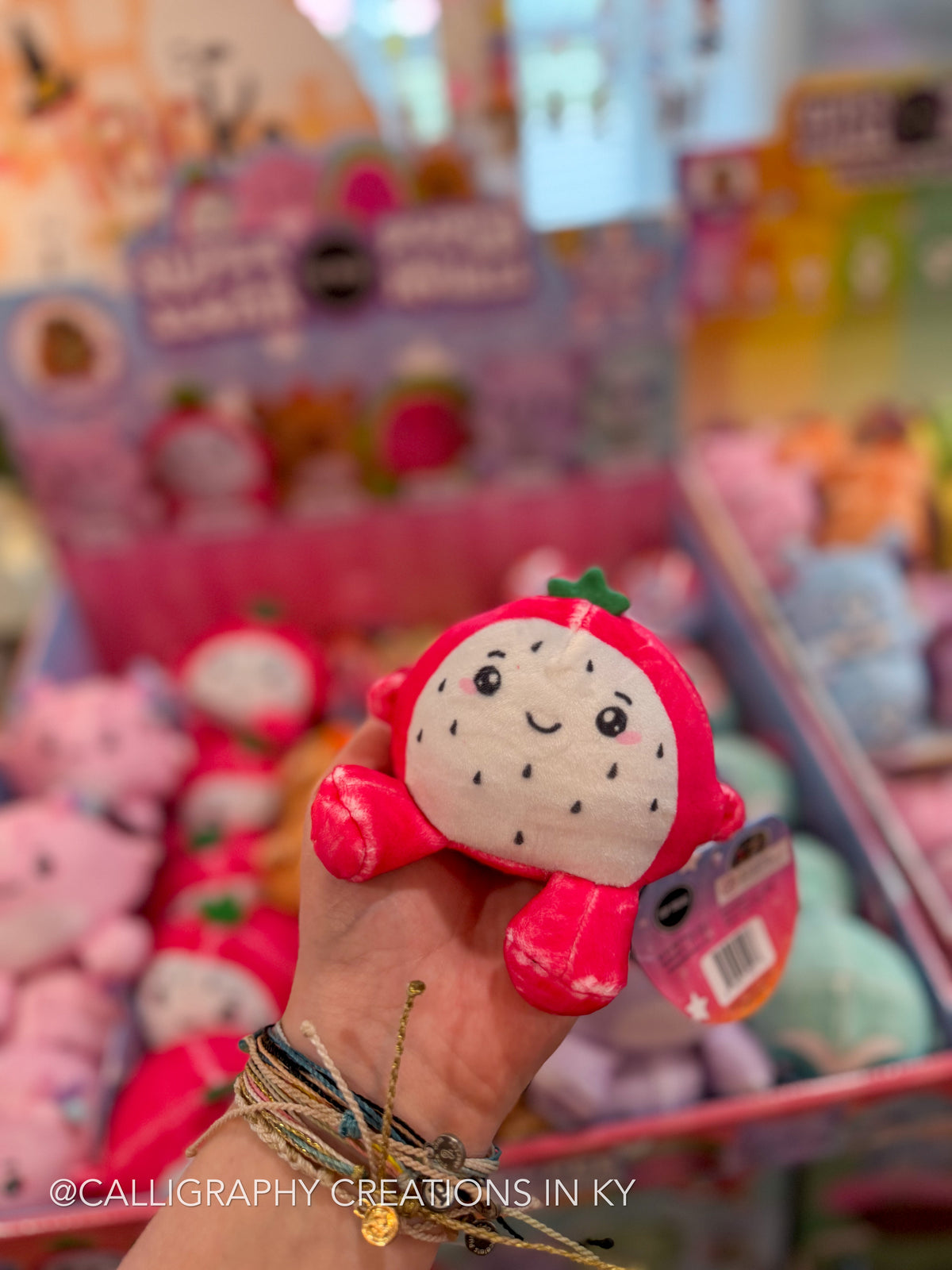 A hand holds a small plush strawberry toy in a brightly lit retail display filled with similar stuffed animals. The toy has a white face with black embroidered seeds and a pink-red body with a green leaf on top.