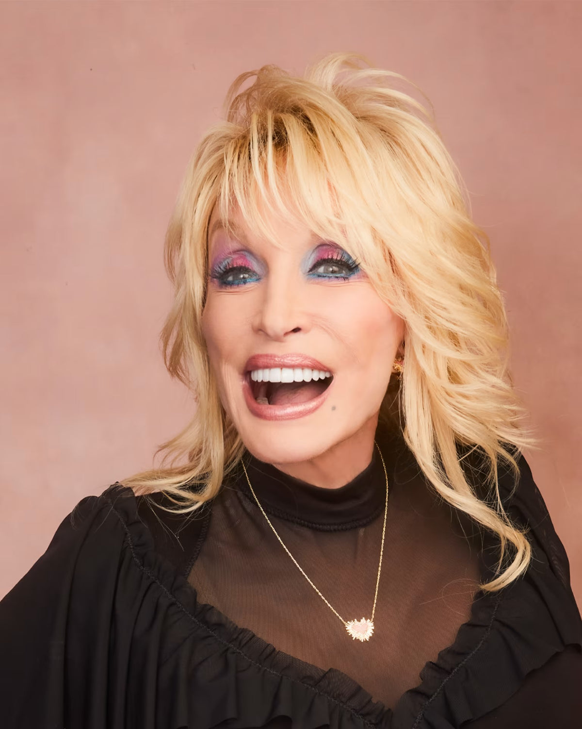 Dolly Parton smiling broadly, showcasing her signature blonde hair, colorful blue and purple eye makeup, and a black sheer ruffled top with a gold necklace, set against a solid dusty pink background.