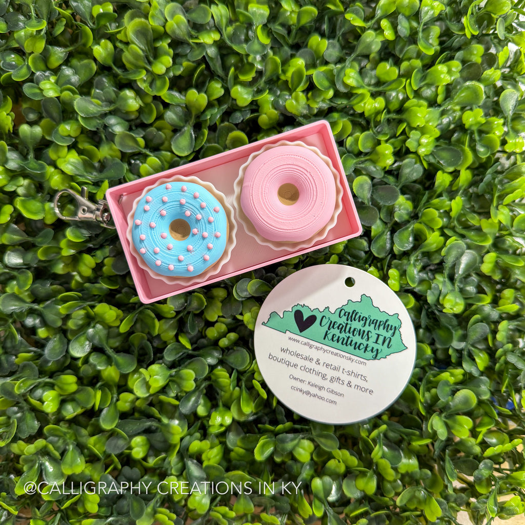 Pink box with donut-shaped fidget clickers in the form of a keychain on a green leafy background, featuring Calligraphy Creations in Kentucky branding.