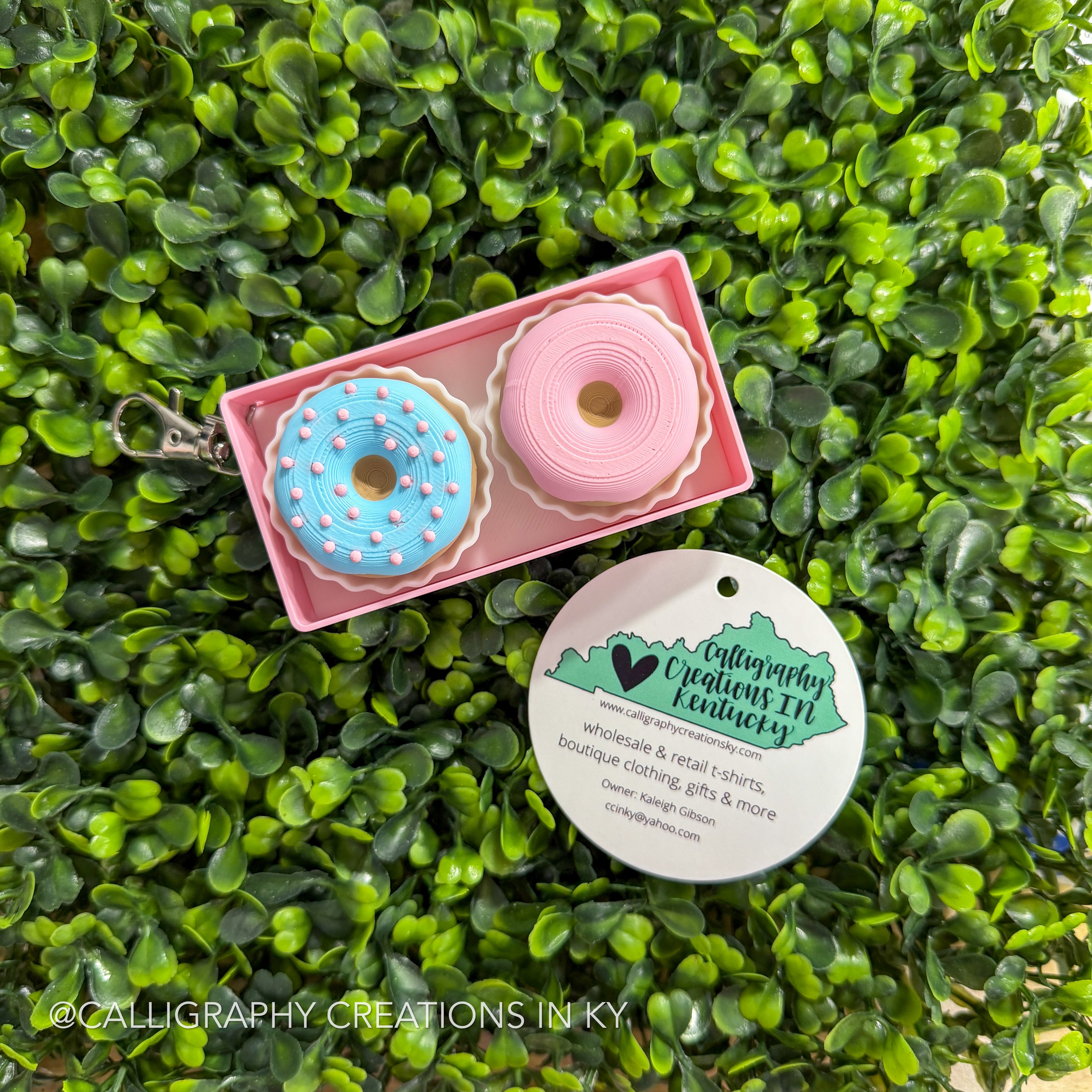 Pink box with donut-shaped fidget clickers in the form of a keychain on a green leafy background, featuring Calligraphy Creations in Kentucky branding.