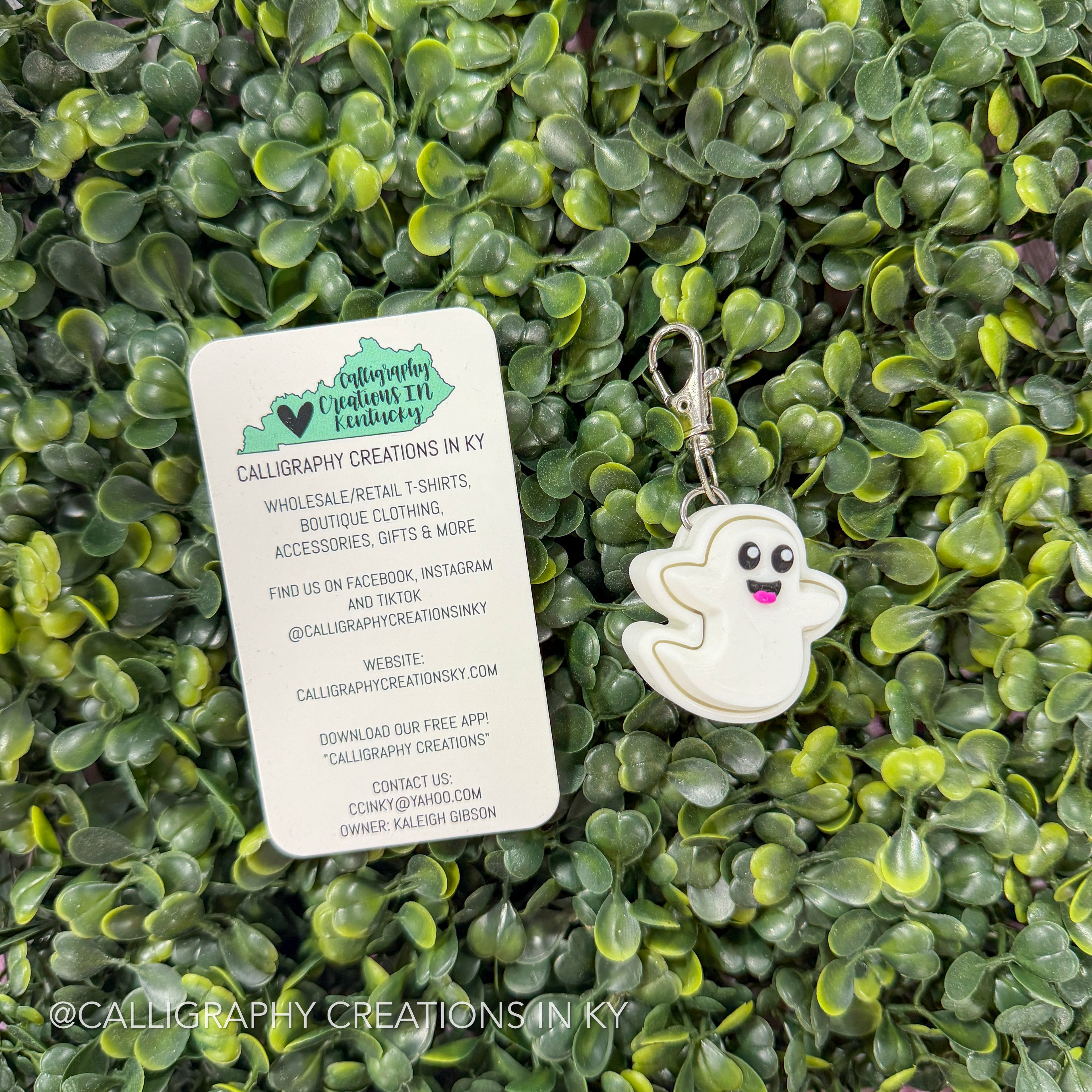 A white ghost-shaped keychain with a silver clasp resting on a white card, placed on a background of green leafy vines. The card shows the brand 'Calligraphy Creations In Ky'.