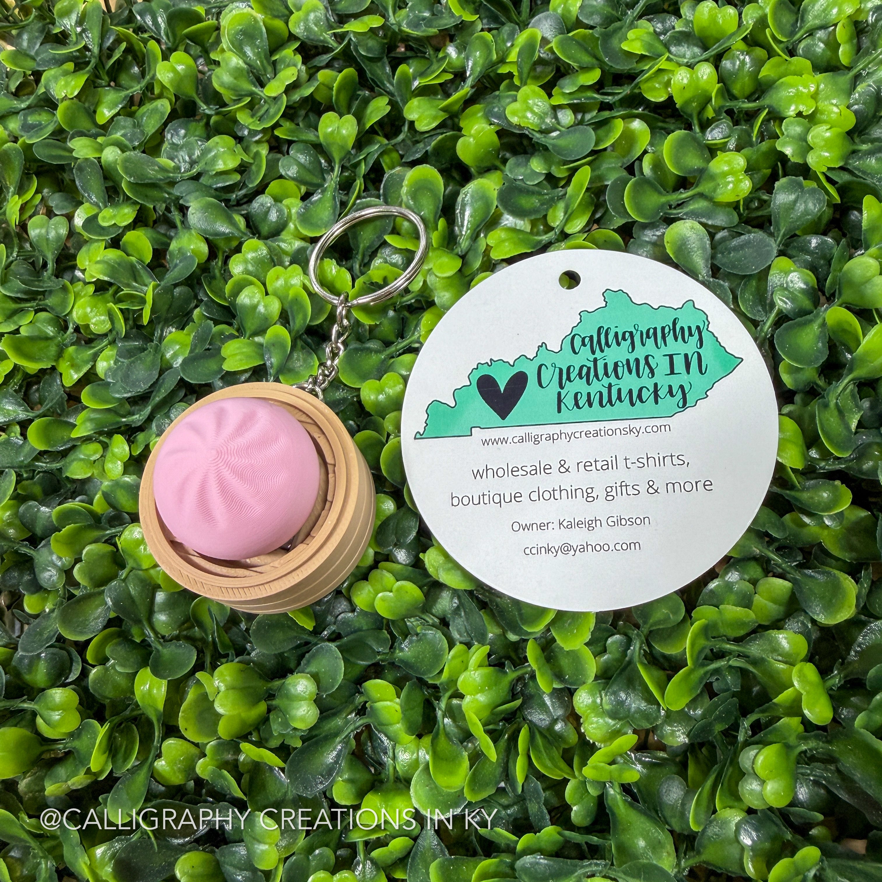 Keychain with pink dumpling clicker and 'Calligraphy Creations IN Kentucky' tag on green foliage