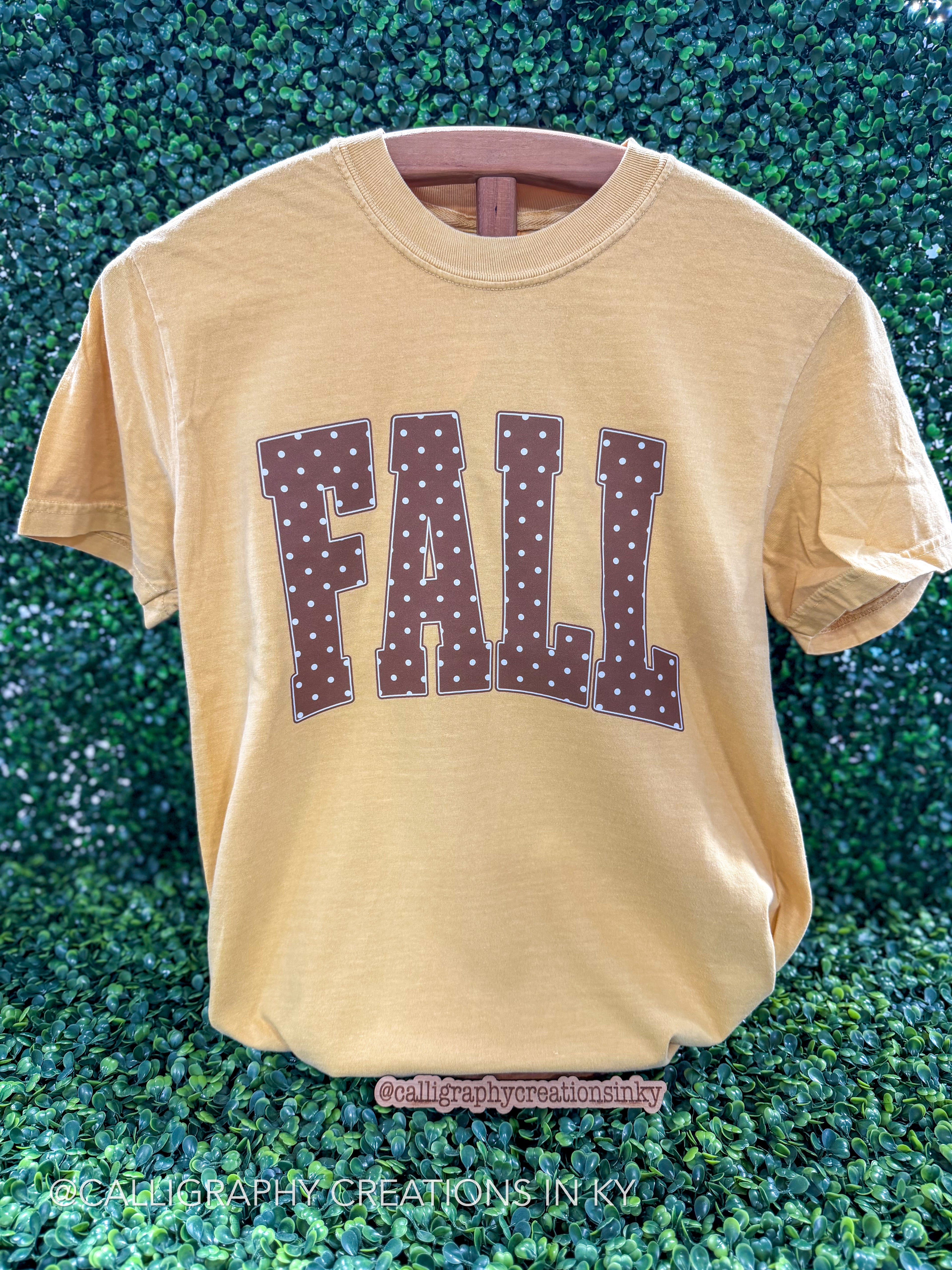 A light tan t-shirt with 'FALL' printed in brown varsity letters with white polka dots, displayed on a wooden hanger against a green hedge background.