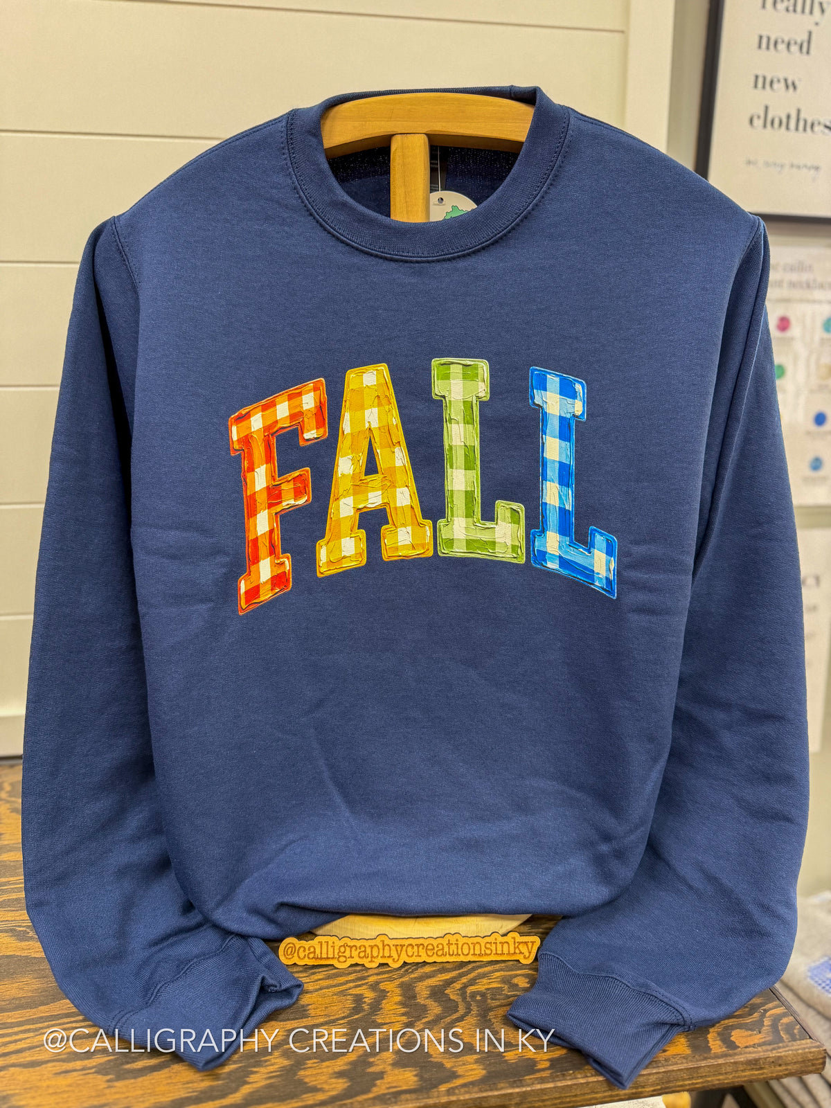 A navy blue crewneck sweatshirt with the word 'FALL' printed across the chest in colorful plaid-style letters (red/orange, yellow, green, blue), hanging on a wooden hanger.