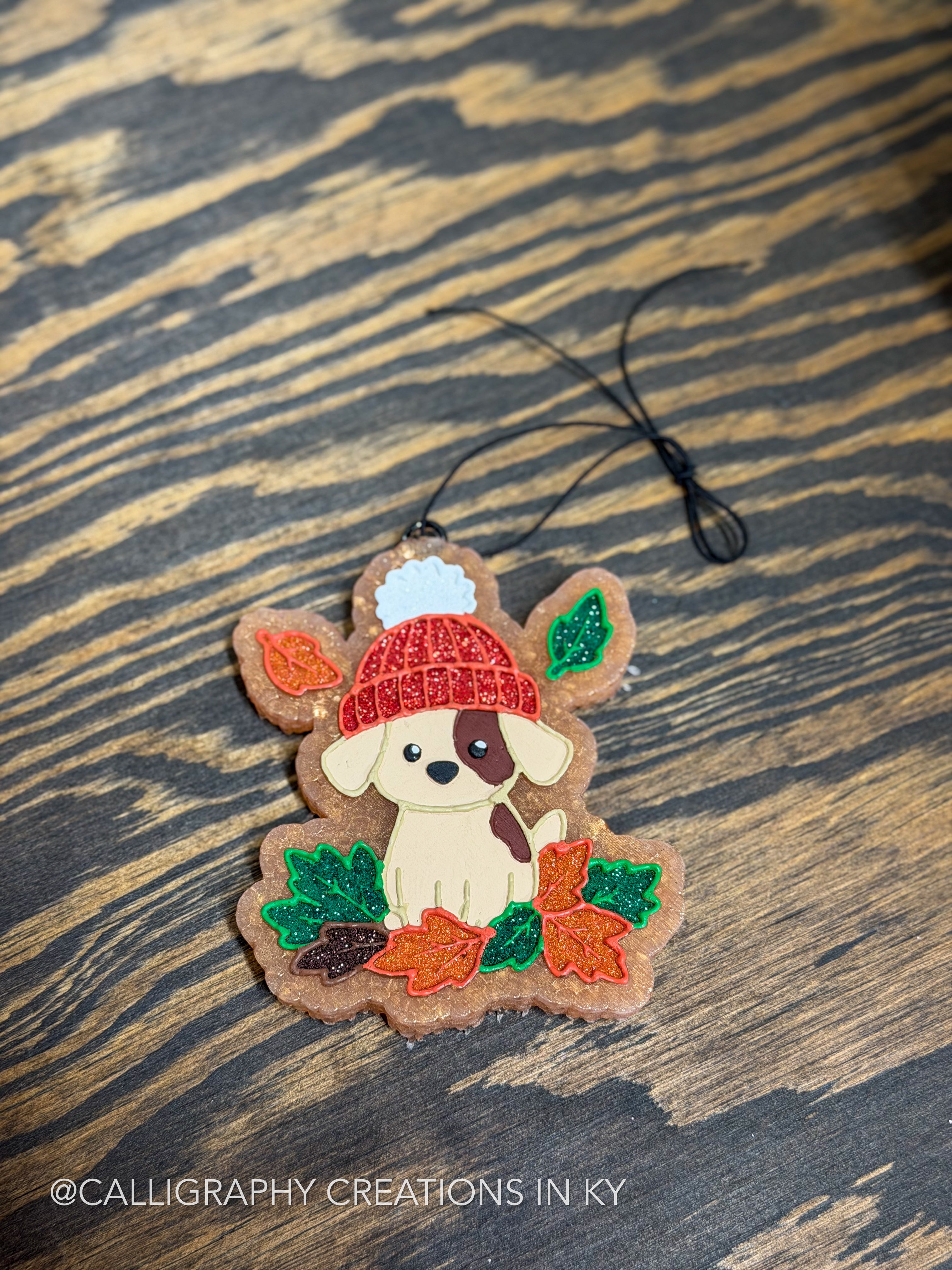 A handmade freshie featuring a cute white puppy wearing a red knit hat and surrounded by autumn leaves, displayed on a dark wood background.