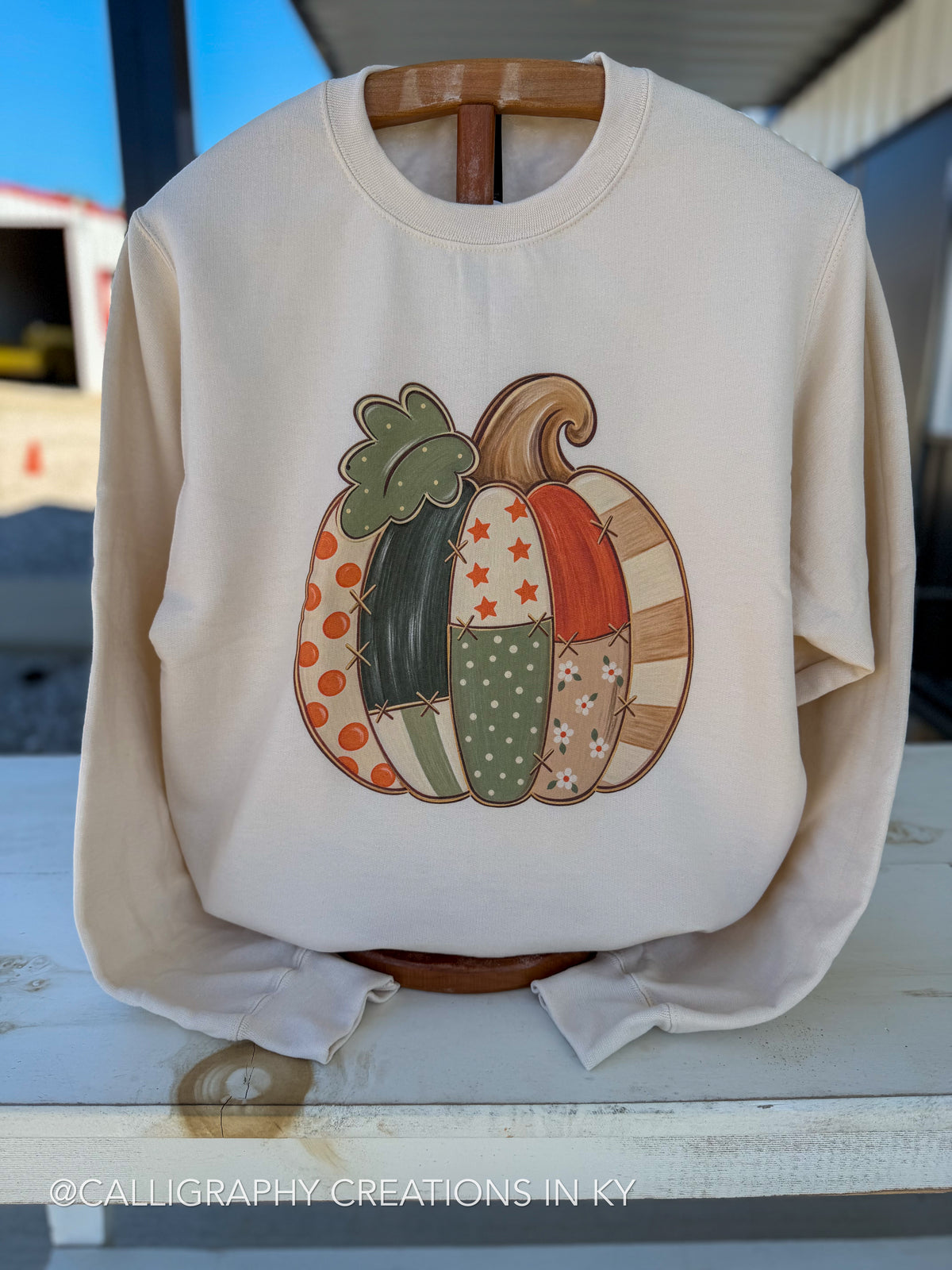 An off-white long-sleeve crewneck sweatshirt displayed on a wooden hanger outdoors. The front features a graphic of three patchwork-style pumpkins in autumn colors (orange, green, brown, beige) with patterns like polka dots, stars, stripes, and leaves.