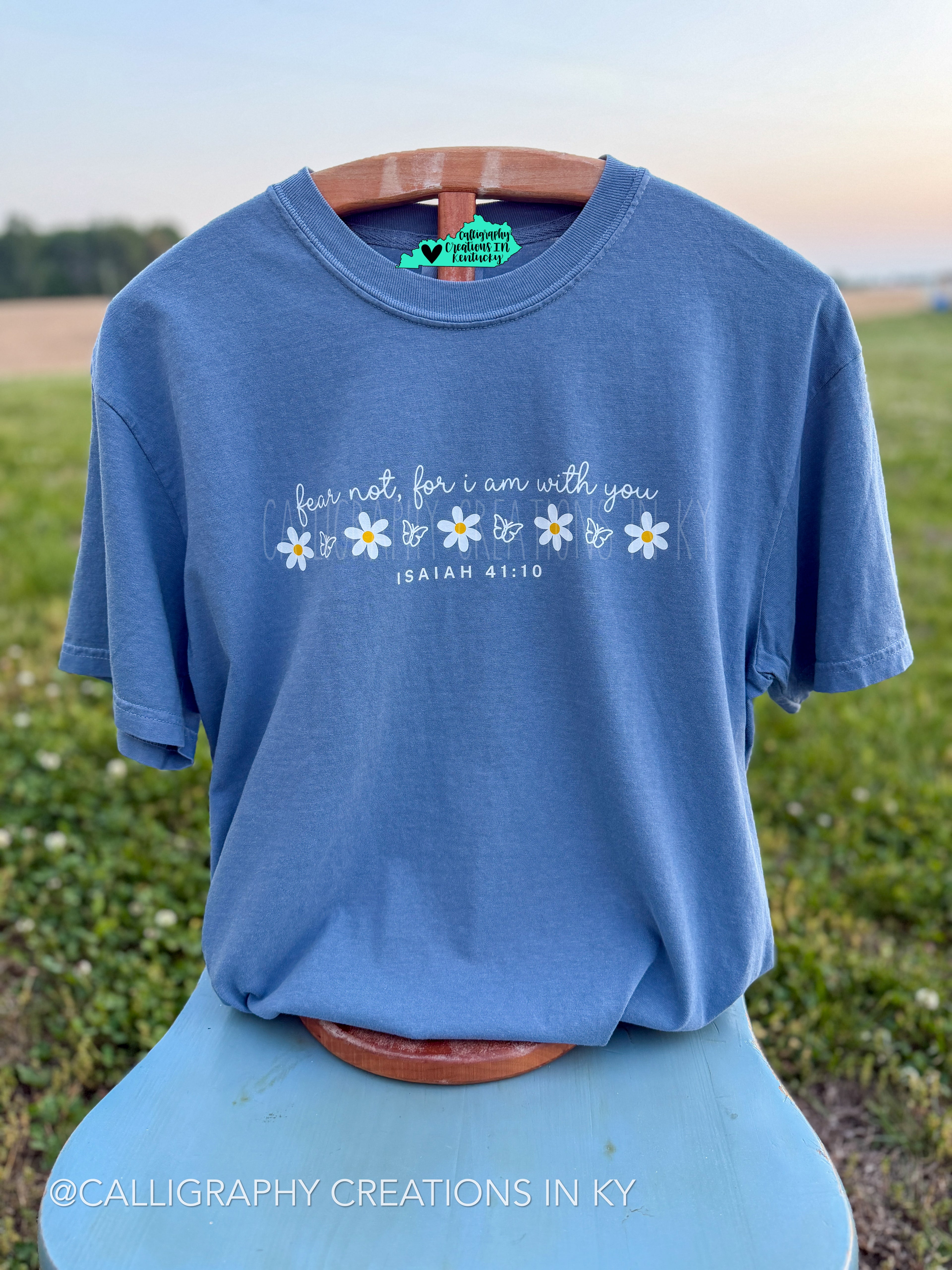 Blue Jean colored comfort colors tee with white lettering on the tee. An open field background