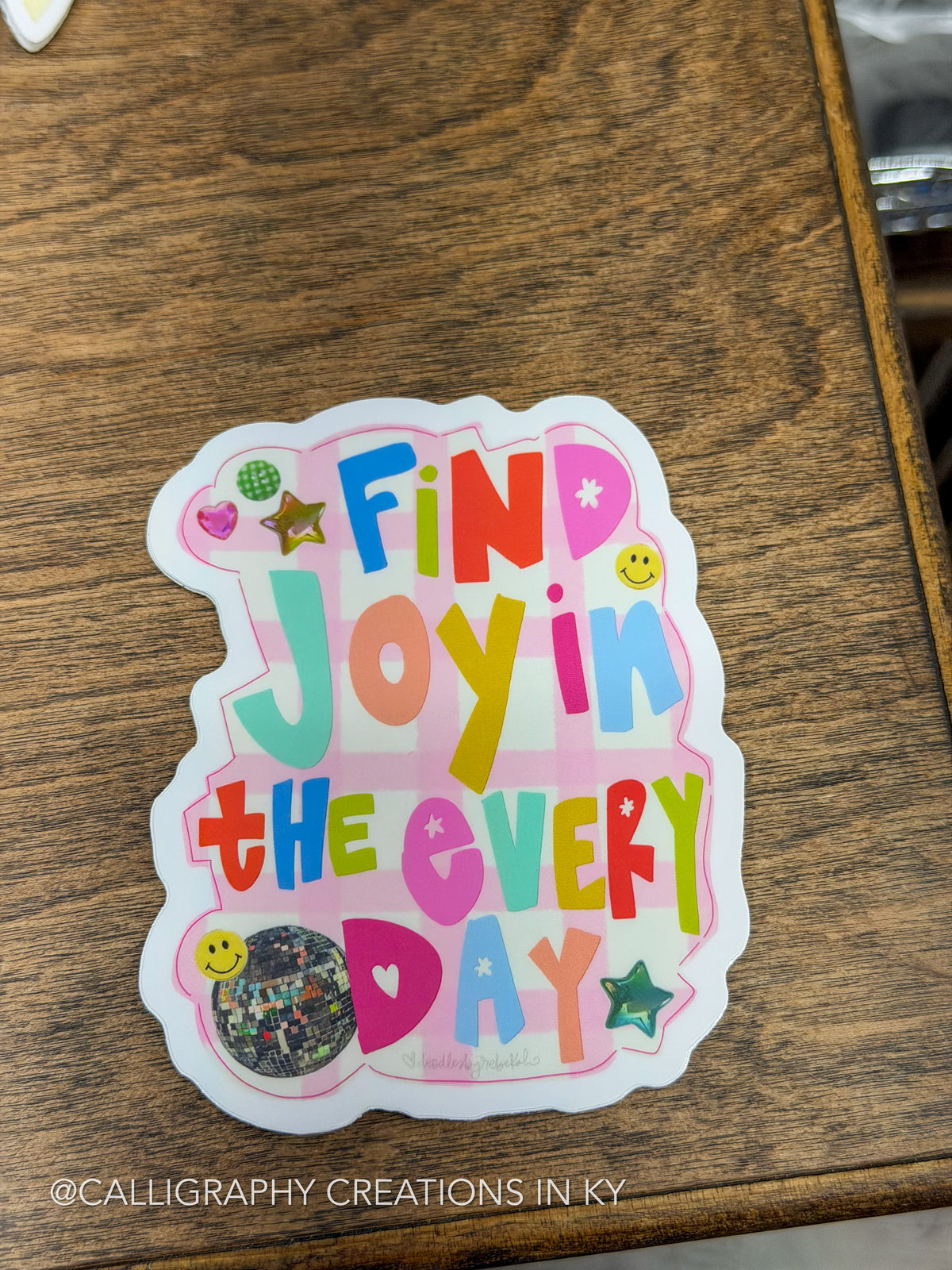 A die-cut sticker with the phrase 'Find Joy in the Every Day' in colorful retro-style lettering, decorated with small smiley faces, stars, hearts, and a glittery circular element, placed on a wooden surface.
