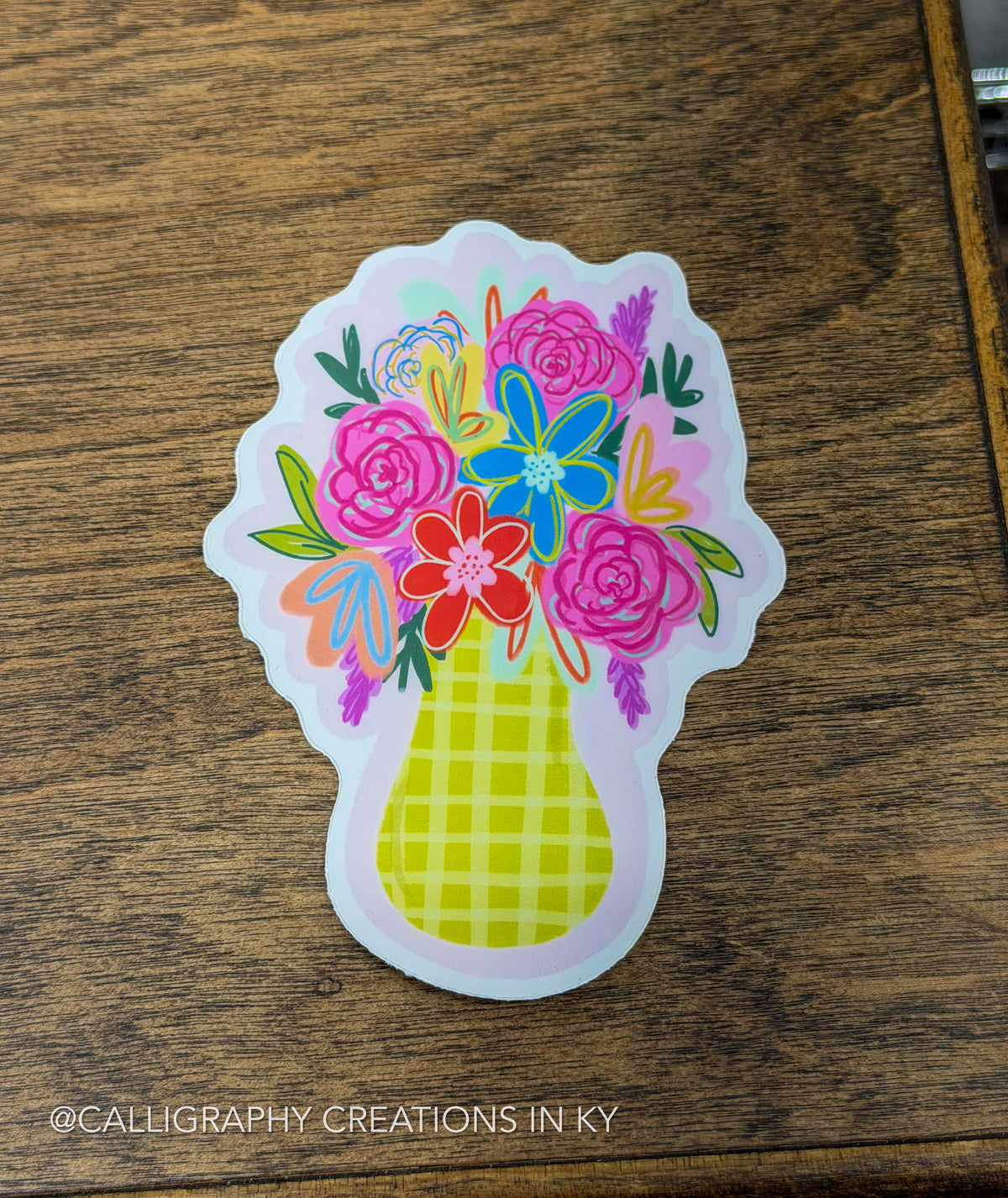 A die-cut sticker featuring a colorful floral bouquet in a yellow-green checkered vase, placed on a dark wood surface. The sticker has a white border and is photographed from above.