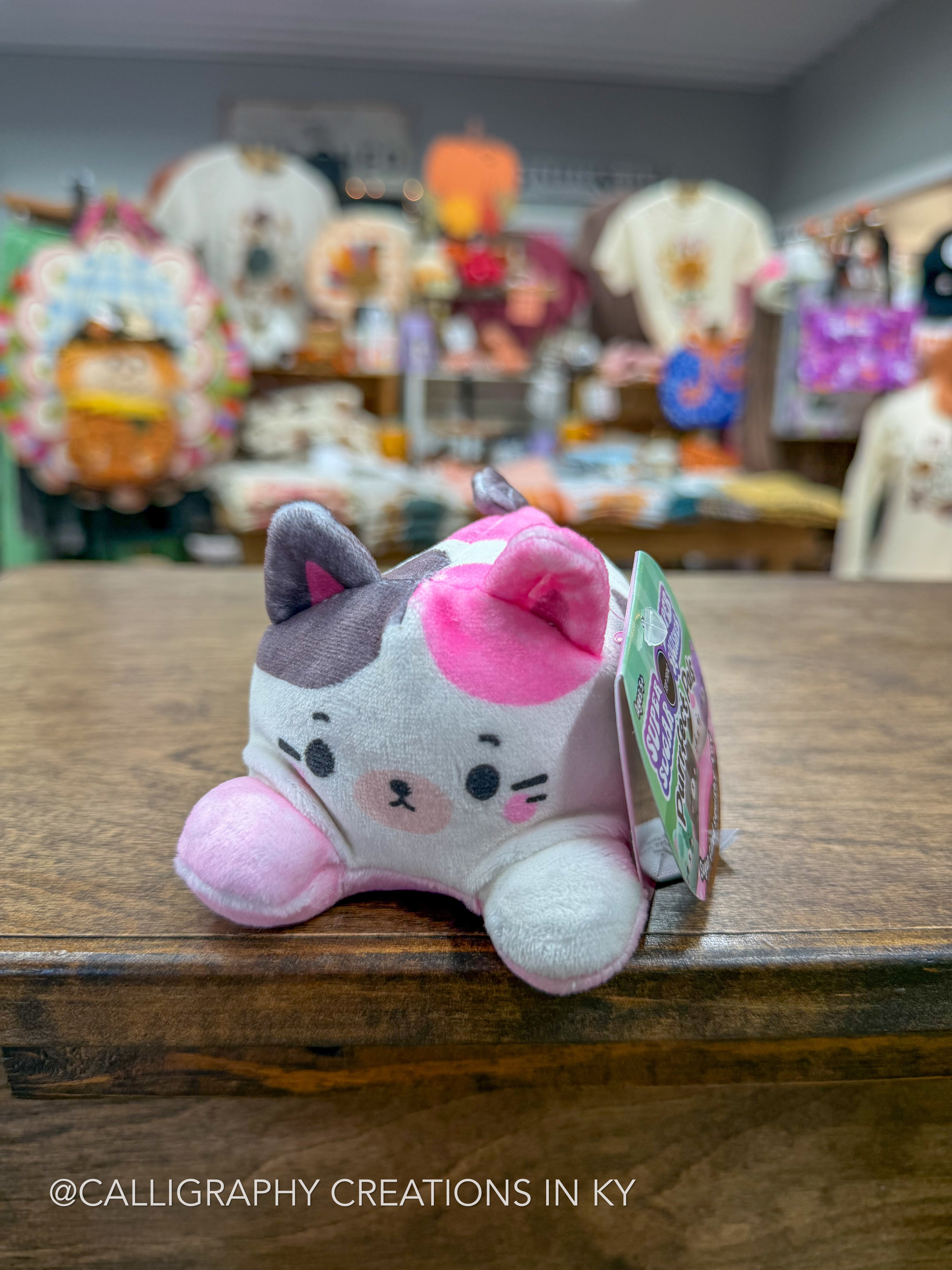 A small plush cat toy with pink and gray patches sits on a wooden counter in a shop, with a hangtag attached and a blurred store interior in the background.
