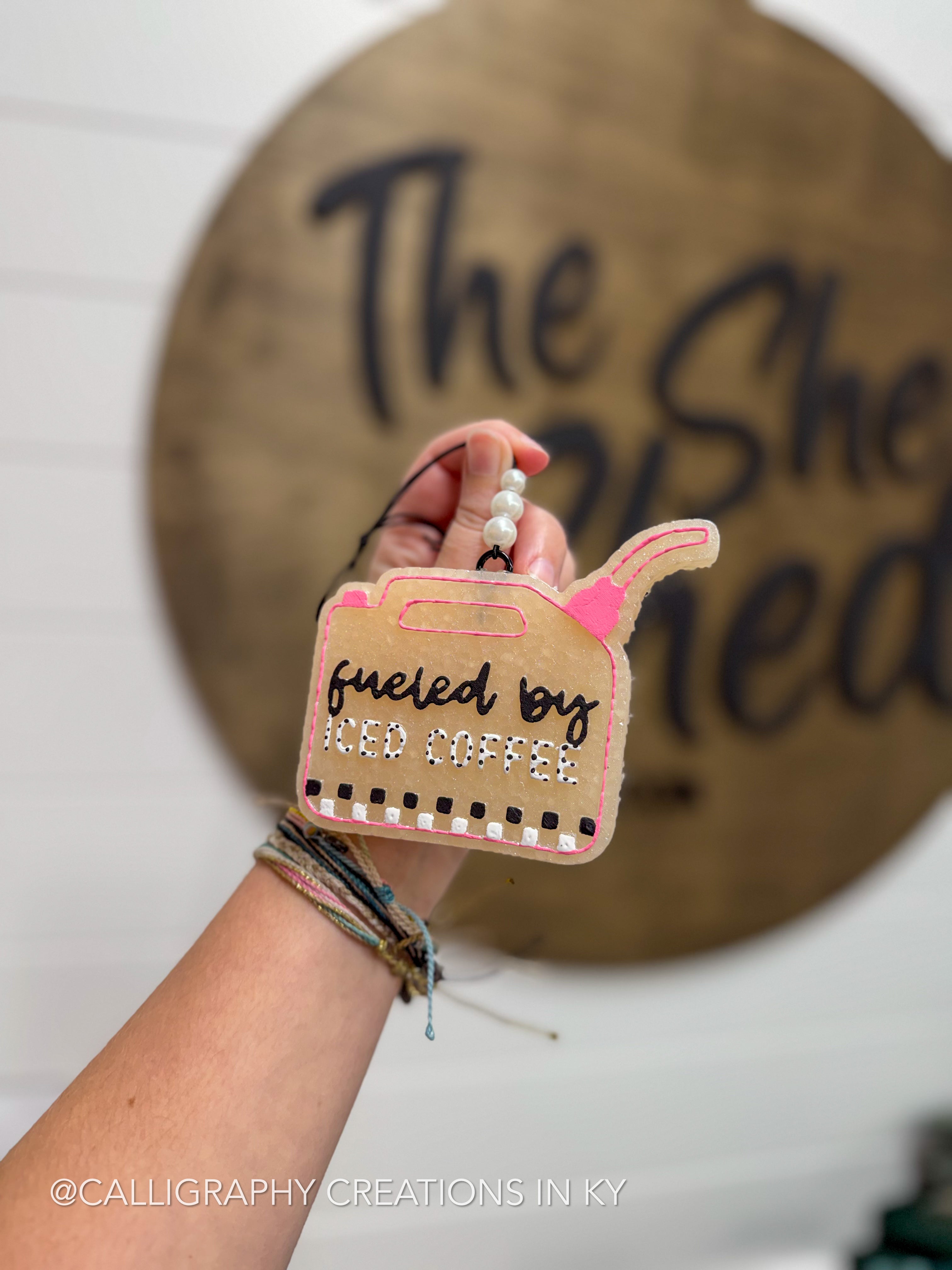 A hand holds a freshie shaped like a fuel gauge, with the text 'fueled by iced coffee' in black script and white rhinestones. The sign has pink accents and is photographed in front of a blurred wooden sign reading 'The She.