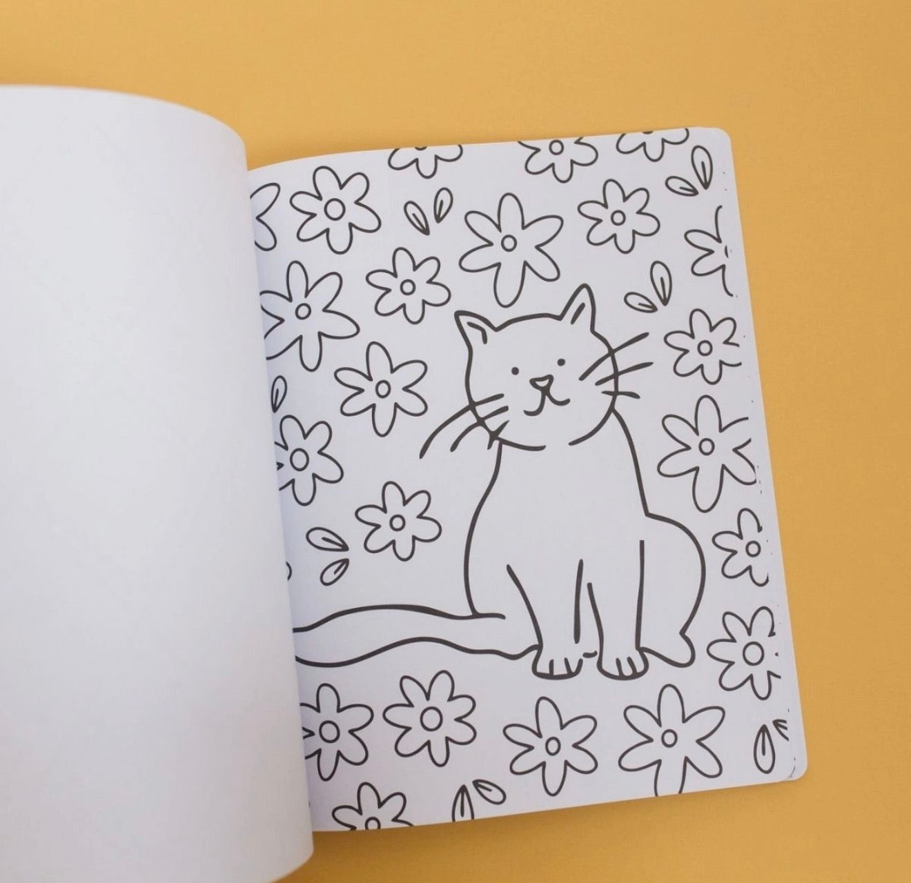 An open notebook or coloring book page with a black line drawing of a cat surrounded by simple flowers on a white page, photographed against a warm yellow background.