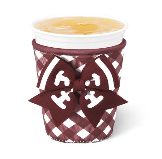 A white paper cup with a yellow beverage, wrapped in a burgundy and white plaid cup sleeve featuring a large matching bow with white lettering.