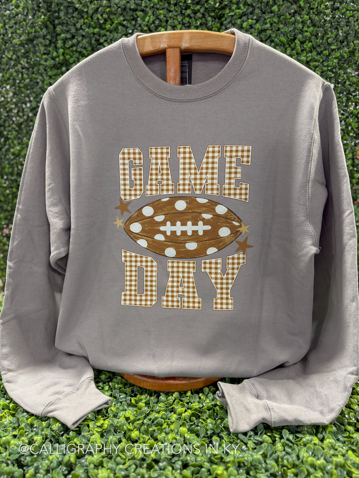 A gray long-sleeve crewneck sweatshirt displayed on a wooden hanger against a green hedge background. The sweatshirt features a 'GAME DAY' football graphic on the front, with 'GAME' and 'DAY' in a brown and beige gingham pattern, and a brown football in the center with white polka dots and laces.