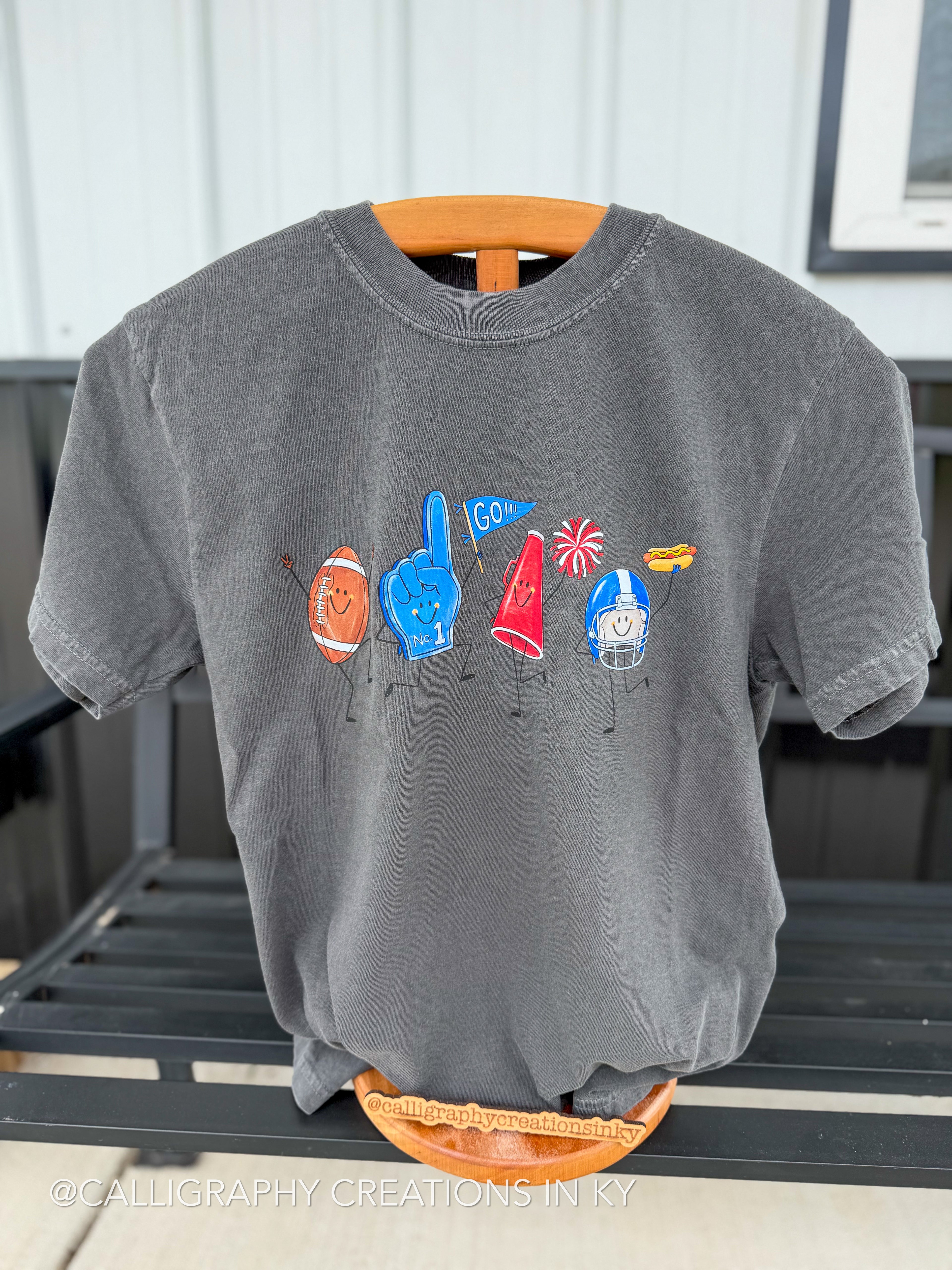 A gray short-sleeve t-shirt on a wooden hanger, featuring a colorful graphic of football-themed characters including an orange football, a blue foam finger, a red megaphone, a blue helmet, and fireworks. The shirt is displayed on a black slatted bench.