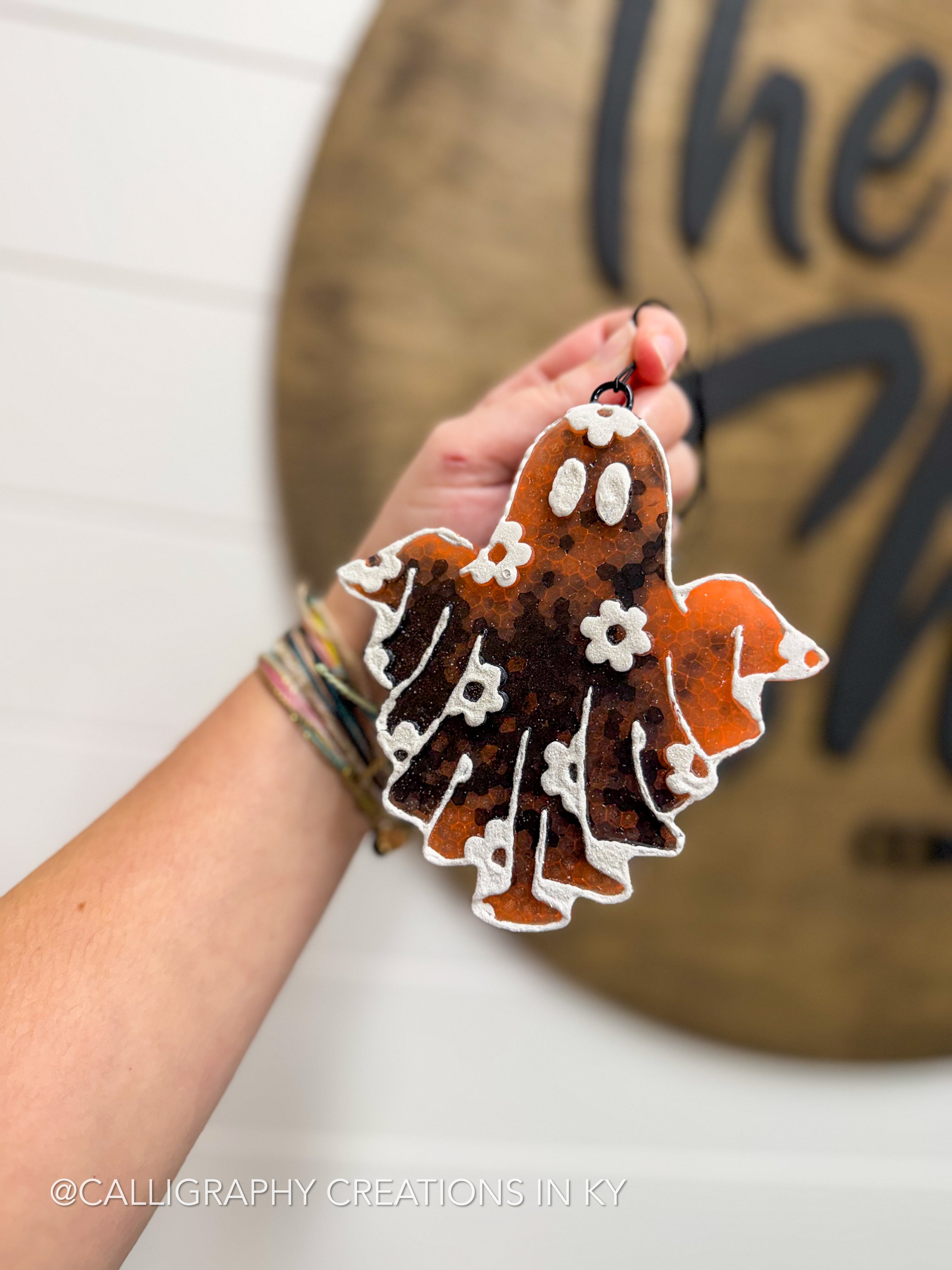 A hand holds a ghost-shaped autumn ornament decorated with orange, brown, and white calligraphic patterns against a blurred background with a wooden sign.