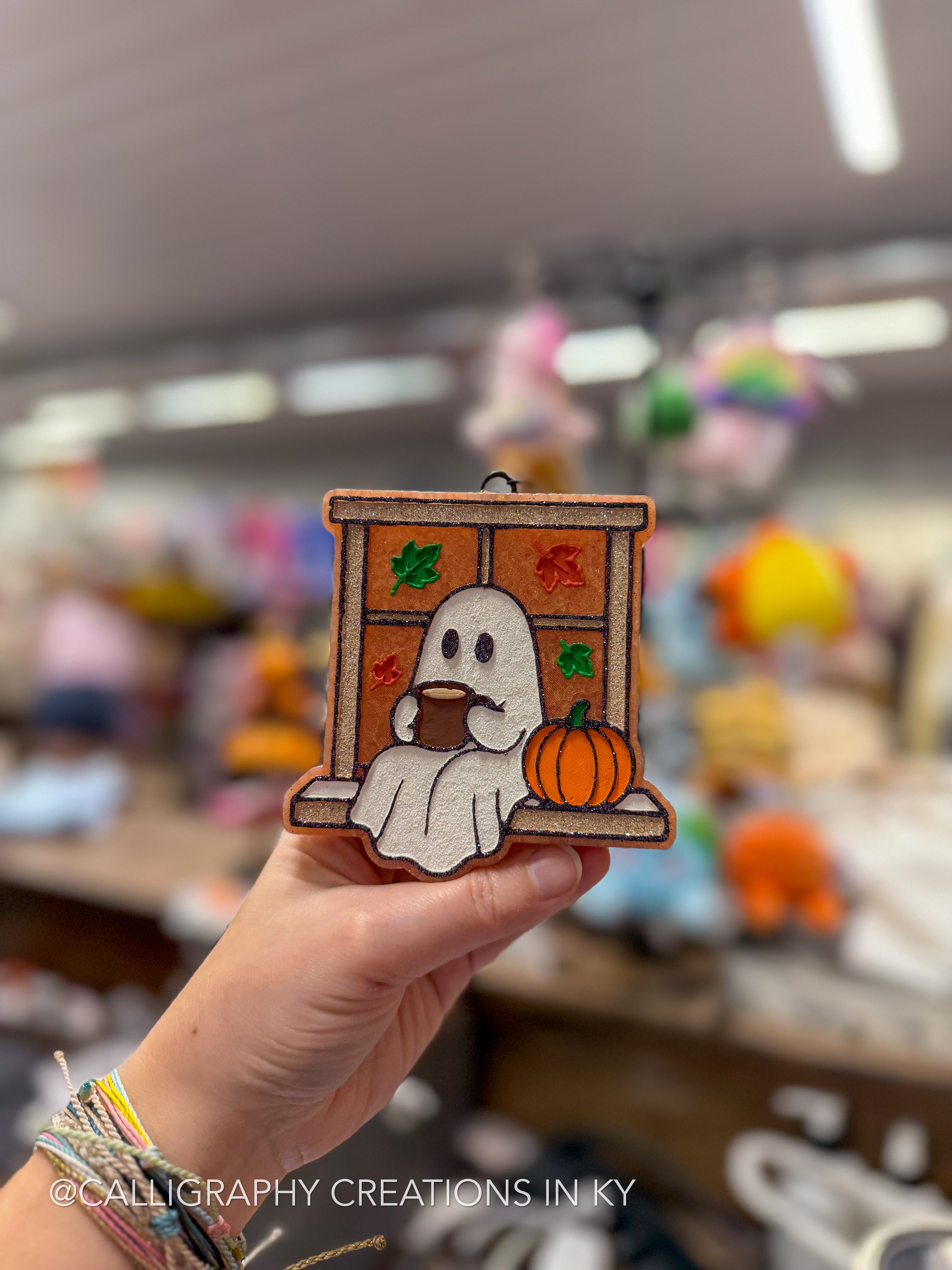 A hand holds a small autumn-themed wooden ornament featuring a ghost at a window with a pumpkin, set against a blurred craft store background.