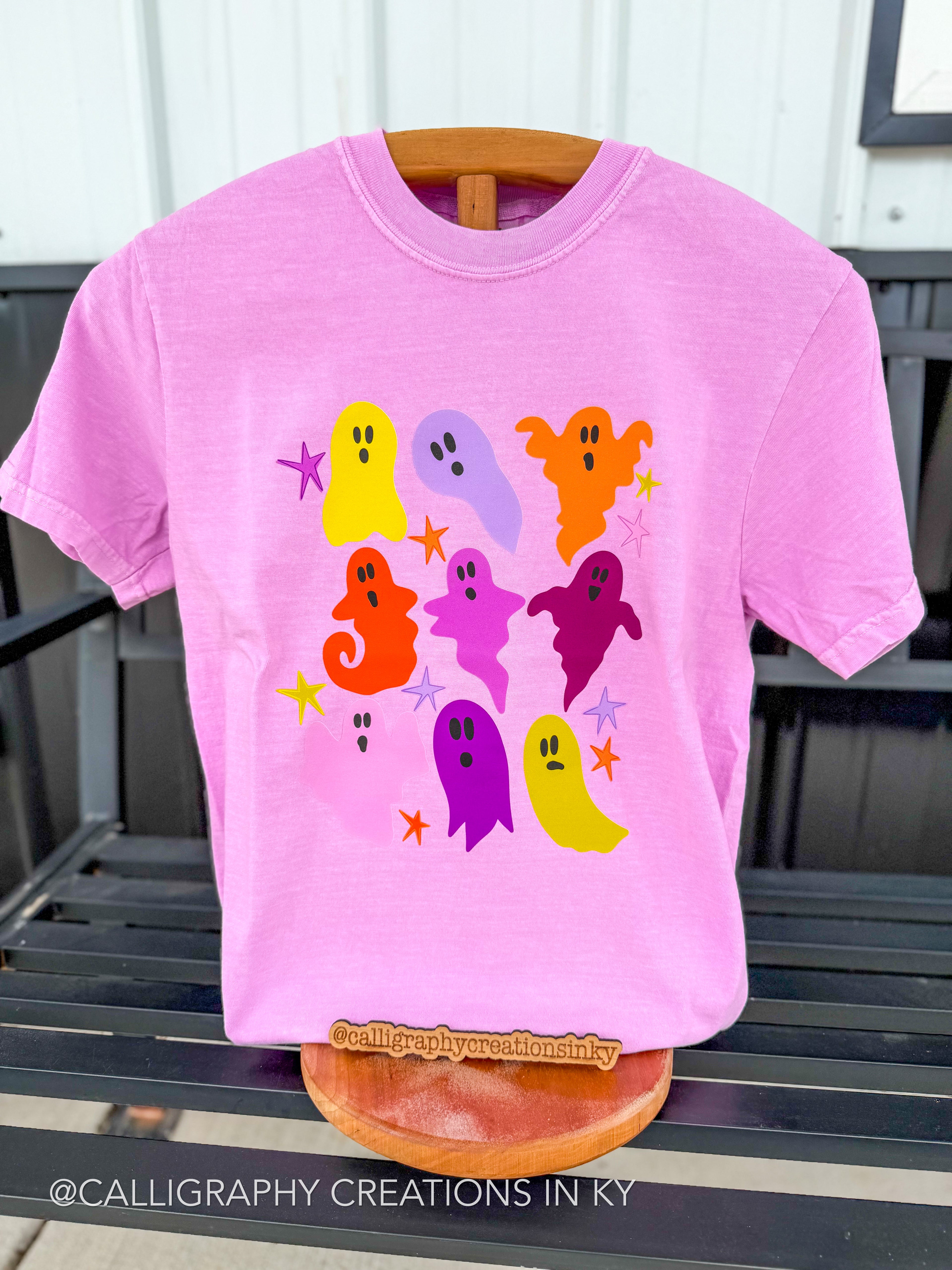 A pink short-sleeve t-shirt displayed on a wooden hanger, featuring a colorful graphic of cute cartoon ghosts in various colors with small star accents. The shirt is photographed on a black slatted bench with a white wall in the background.