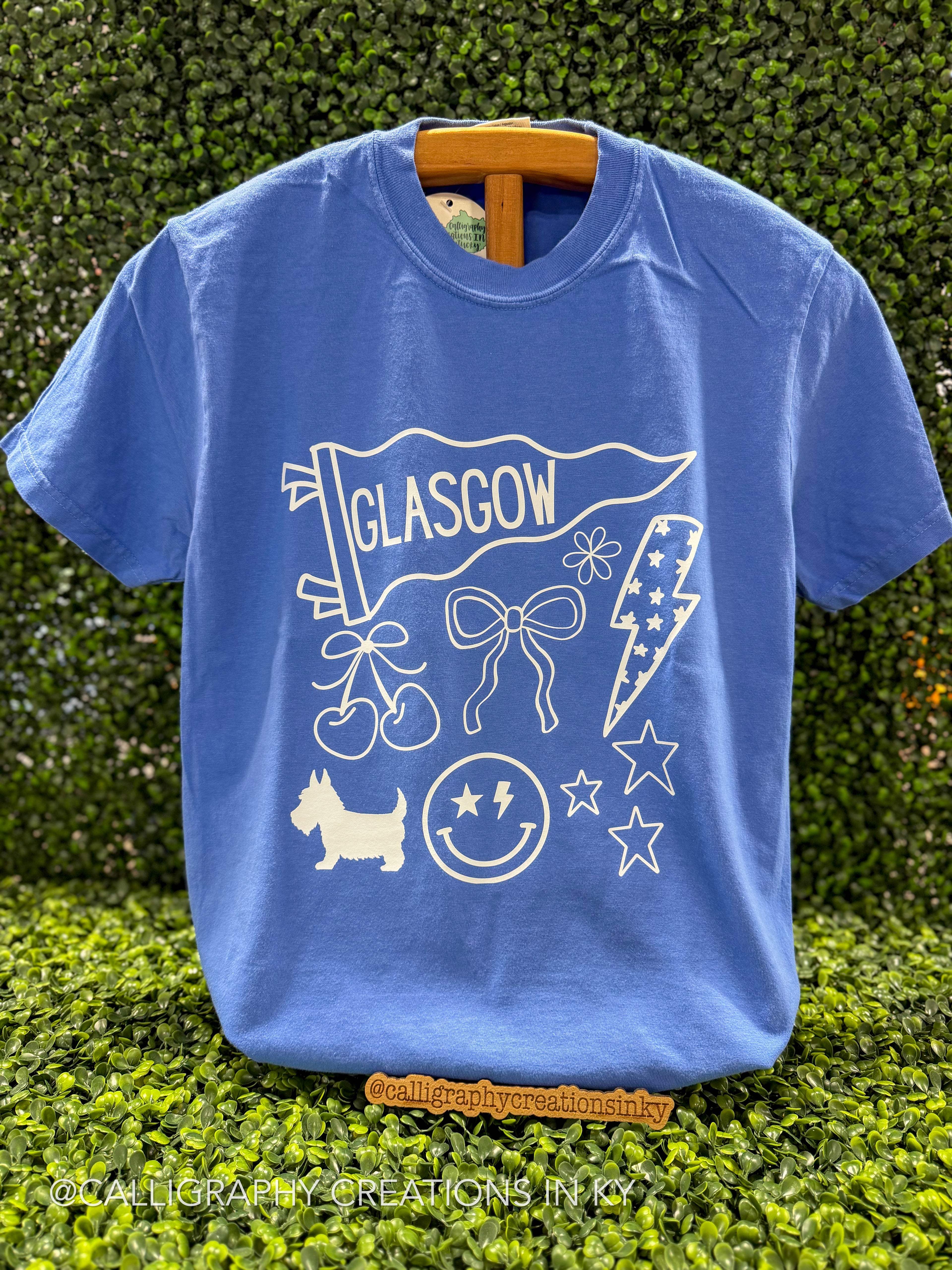 A blue short-sleeve graphic T-shirt hanging on a wooden hanger against a green hedge background. The shirt features a white line-art design with the word 'GLASGOW' on a banner, cherries, a bow, a lightning bolt, stars, a dog silhouette, and a smiley face with stars.