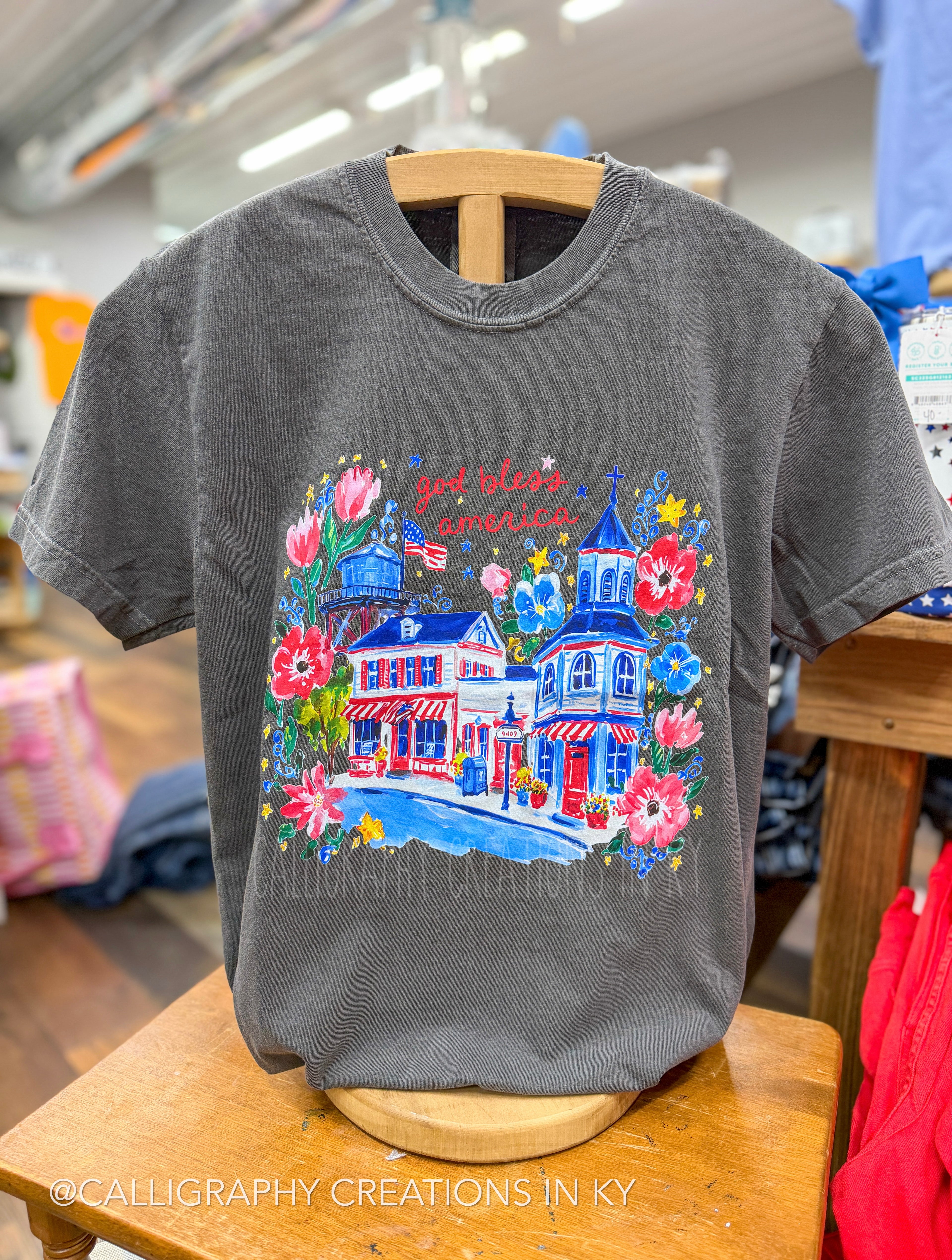 Gray t-shirt with colorful USA graphic design on a wooden hanger in a store setting.