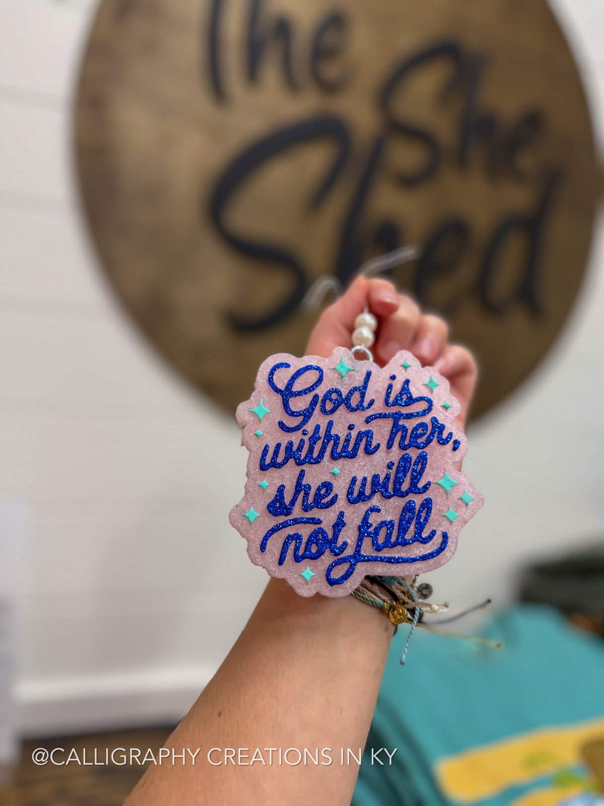 A hand holds a pink freshie with blue cursive text reading 'God is within her, she will not fall' and small blue star accents. The background is a real indoor setting with a blurred wooden sign and wall, making this a Lifestyle image.