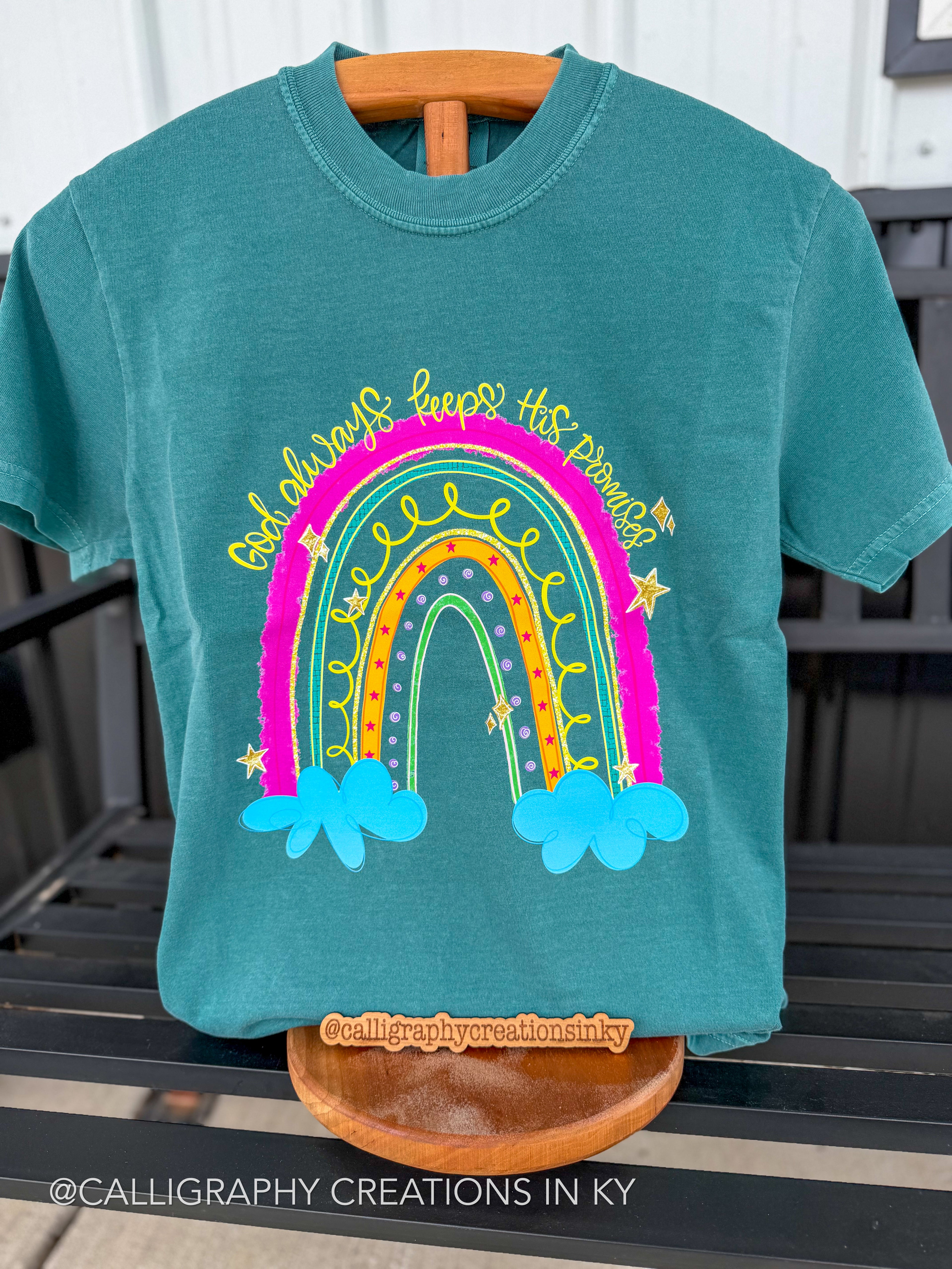 A teal short-sleeve t-shirt displayed on a wooden hanger and stand, featuring a colorful rainbow graphic with the text “God always keeps his promises' in yellow script. The background shows a black slatted outdoor bench.
