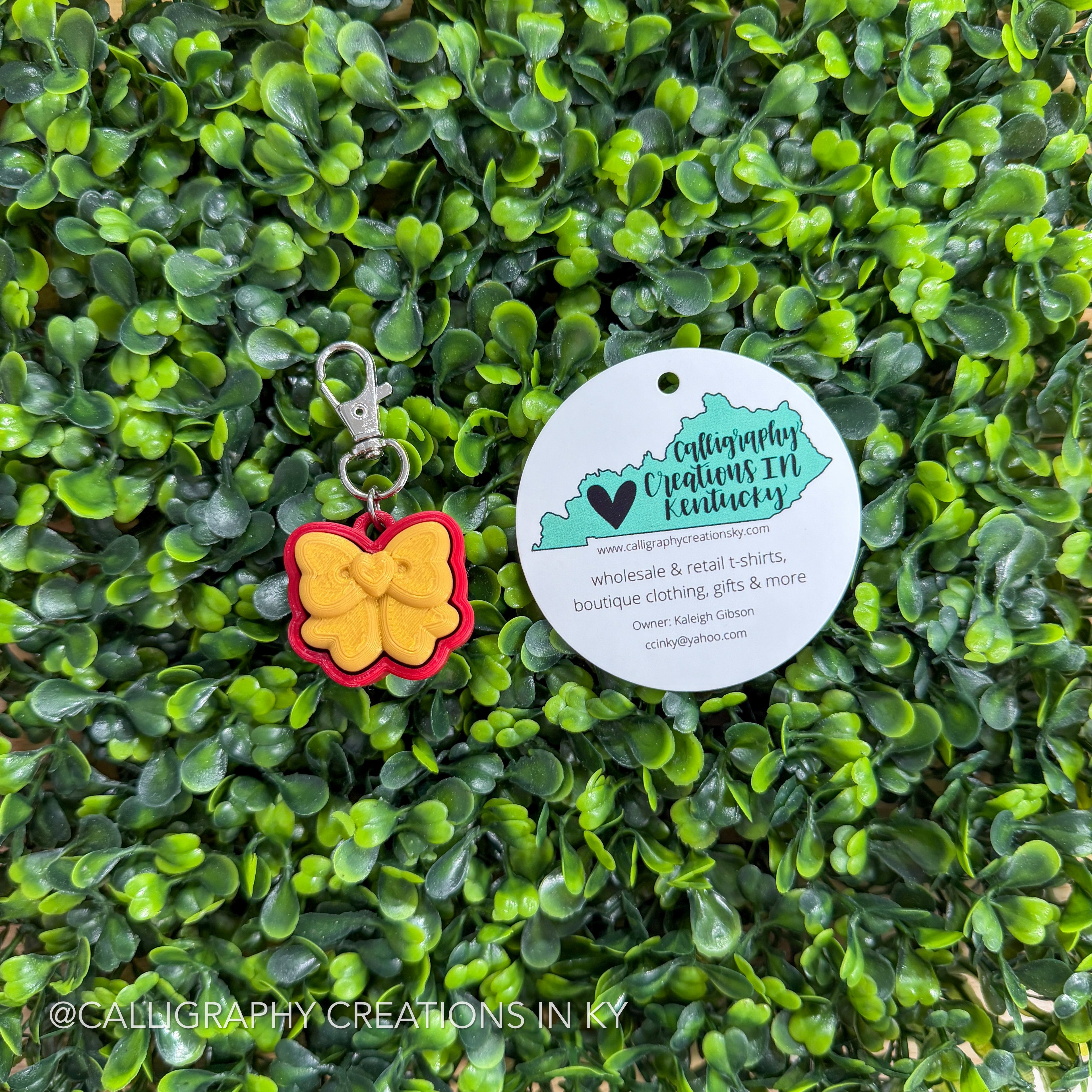 Bow-shaped fidget clicker keychain with a tag on a green leafy background