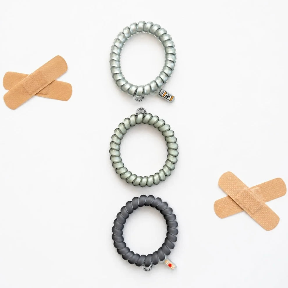 Three coiled elastic wristbands in silver, gray, and black are arranged vertically on a white background, flanked by two beige band-aid props.