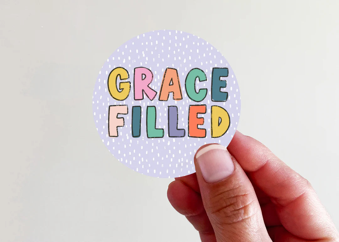 A hand holding a round sticker that reads 'GRACE FILLED' in colorful bubble letters on a light lavender dotted background, set against a plain off-white wall.