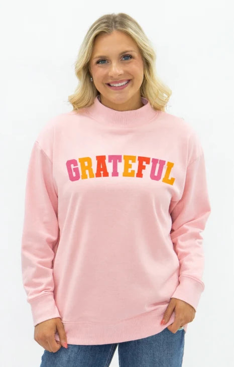 A smiling blonde woman wearing a light pink mock-neck sweatshirt with the word 'GRATEFUL' printed across the chest in colorful gradient letters, paired with blue jeans against a plain white background.