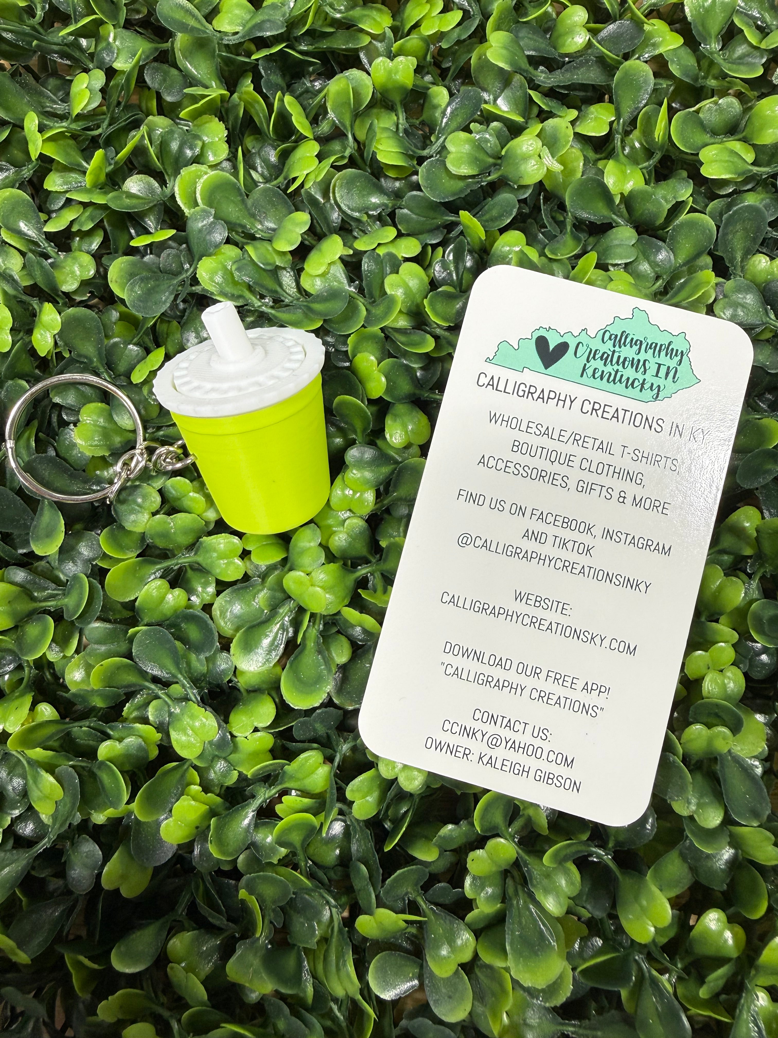 A small green keychain shaped like a miniature cup with a white straw and a silver split ring, placed on a bed of green artificial leaves. A white paper tag with the brand name 'Caligraphy Creations' is resting on top.