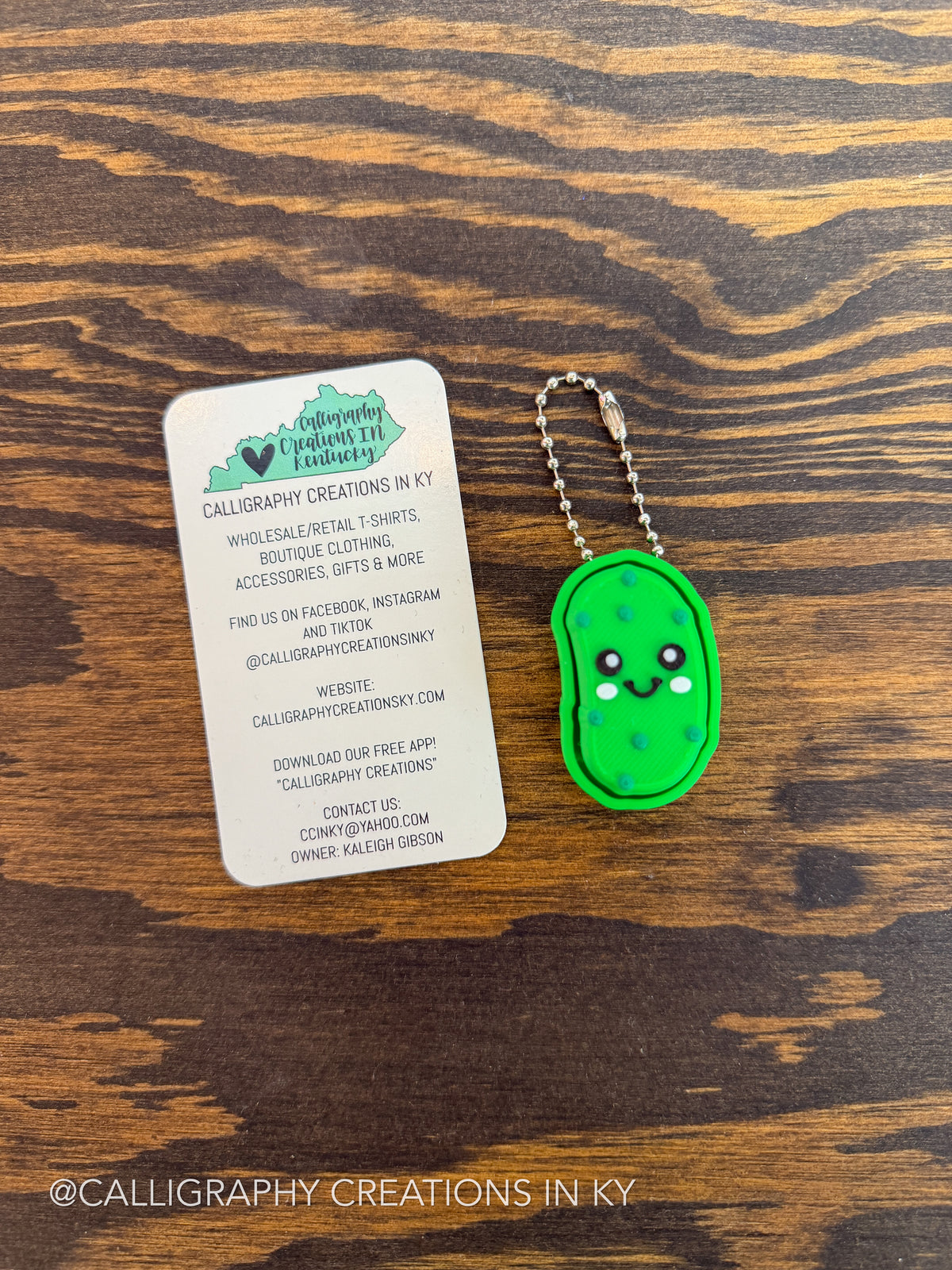 A small green pickle-shaped keychain with a silver ball chain lies next to a white business card on a dark wood surface. The card reads Calligraphy Creations in KY.